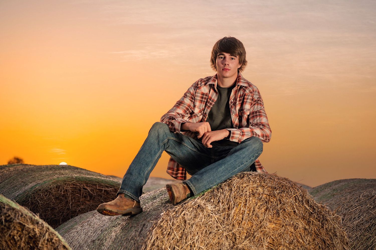 A young man in a plaid shirt and jeans sits on hay bales during a golden sunset in a rural countryside setting.