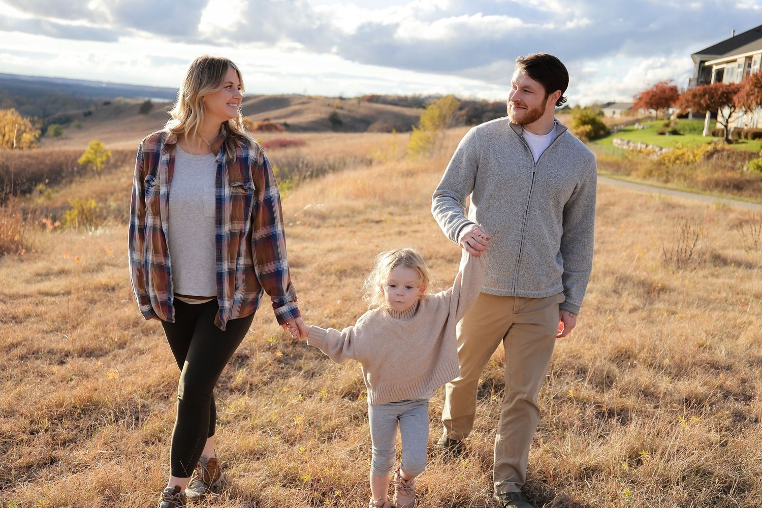 A young family walks hand in hand through a golden autumn hillside meadow.