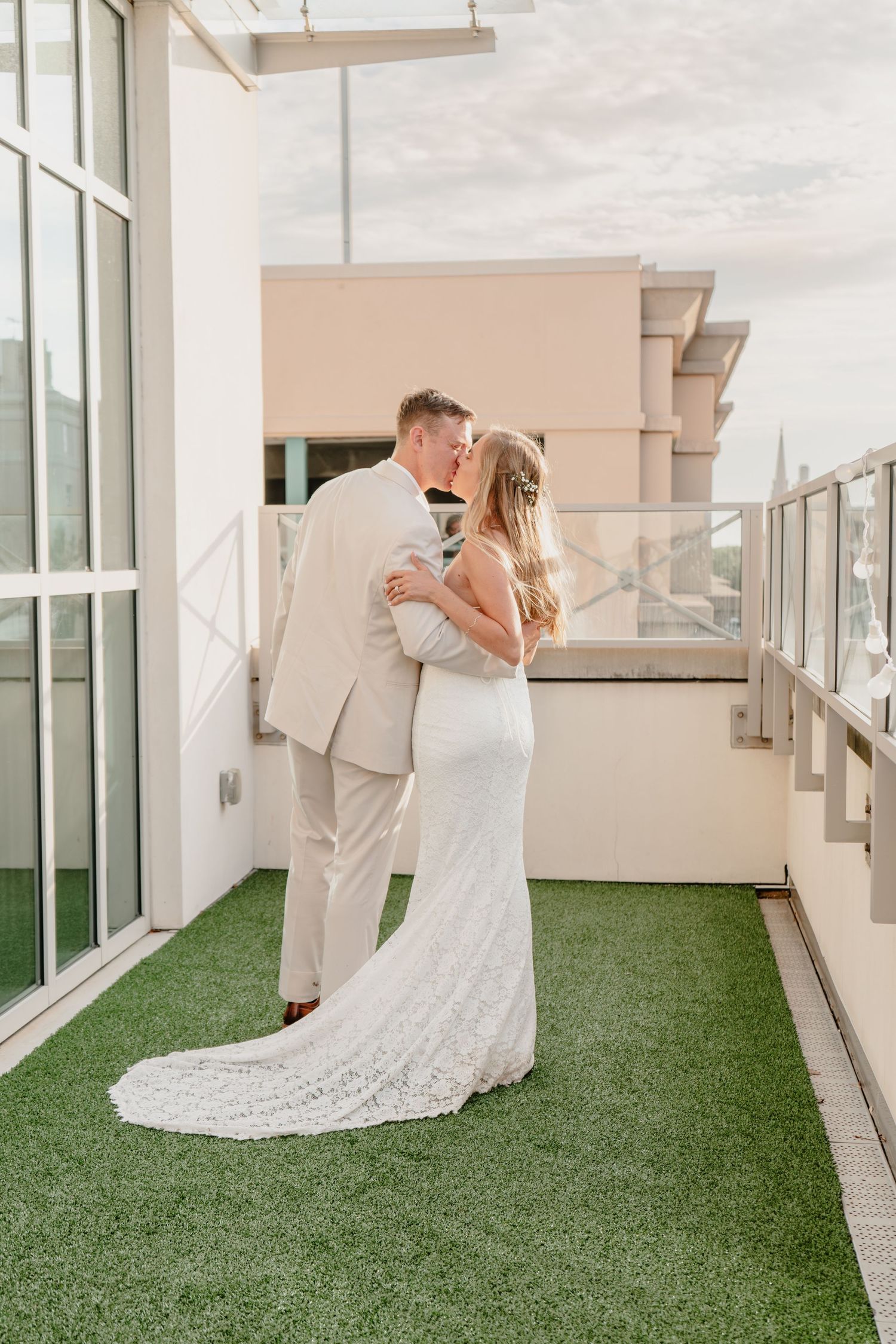 A bride and groom share a romantic kiss on a rooftop terrace, she in a lace gown, he in a tan suit.