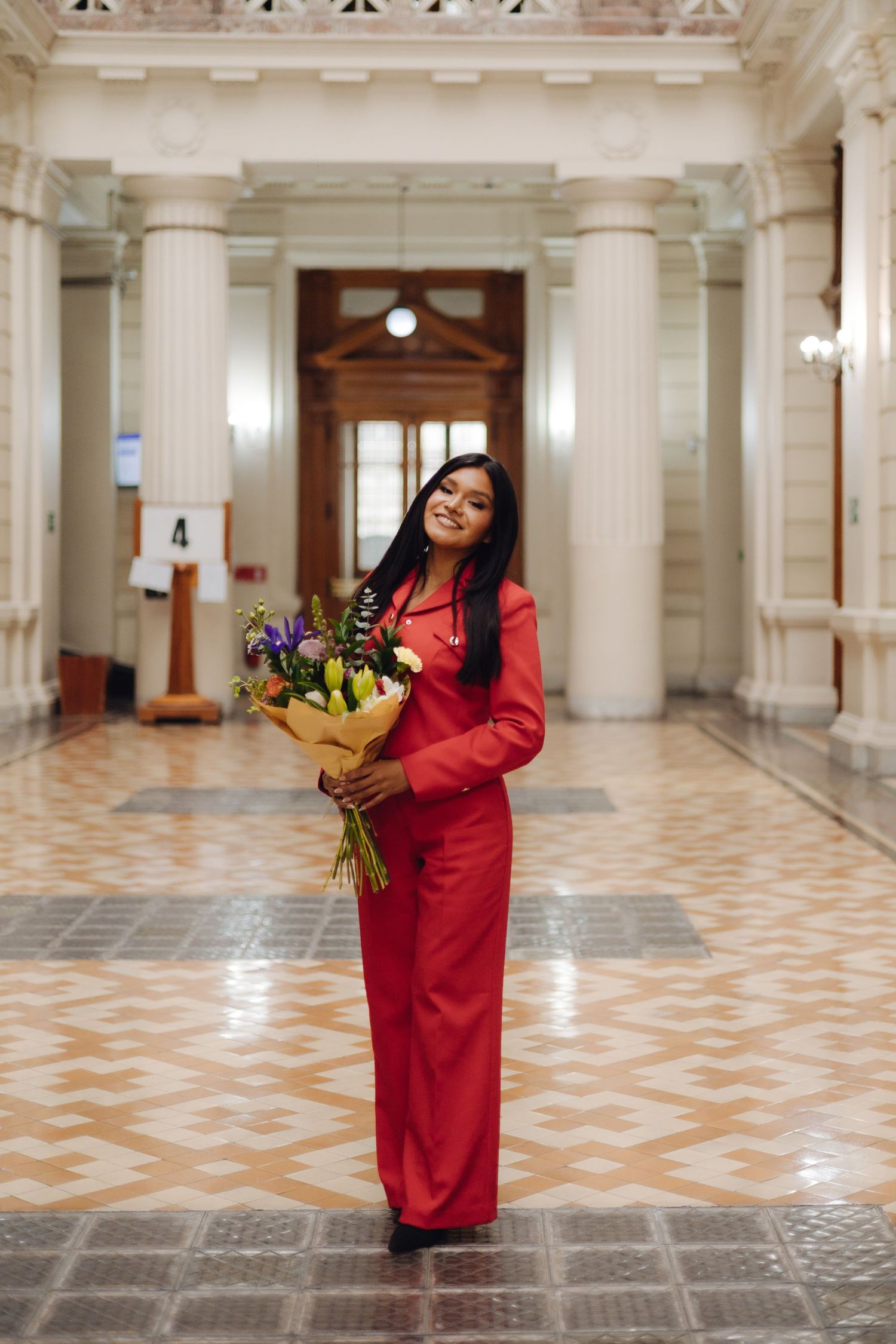 Young woman in red suit holding colorful flower bouquet inside elegant hall.