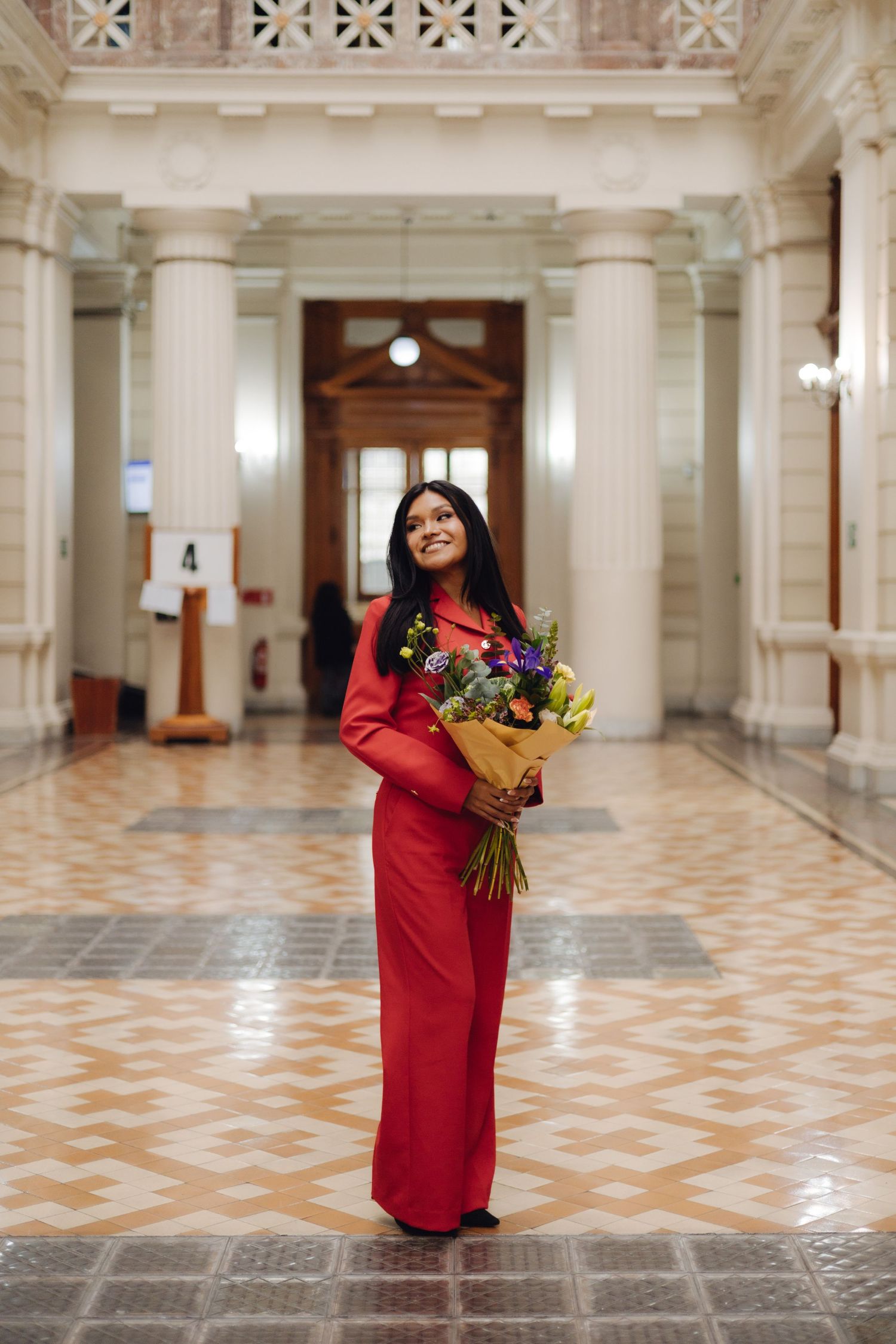 Smiling woman in red outfit holding flowers in grand classical interior hall.
