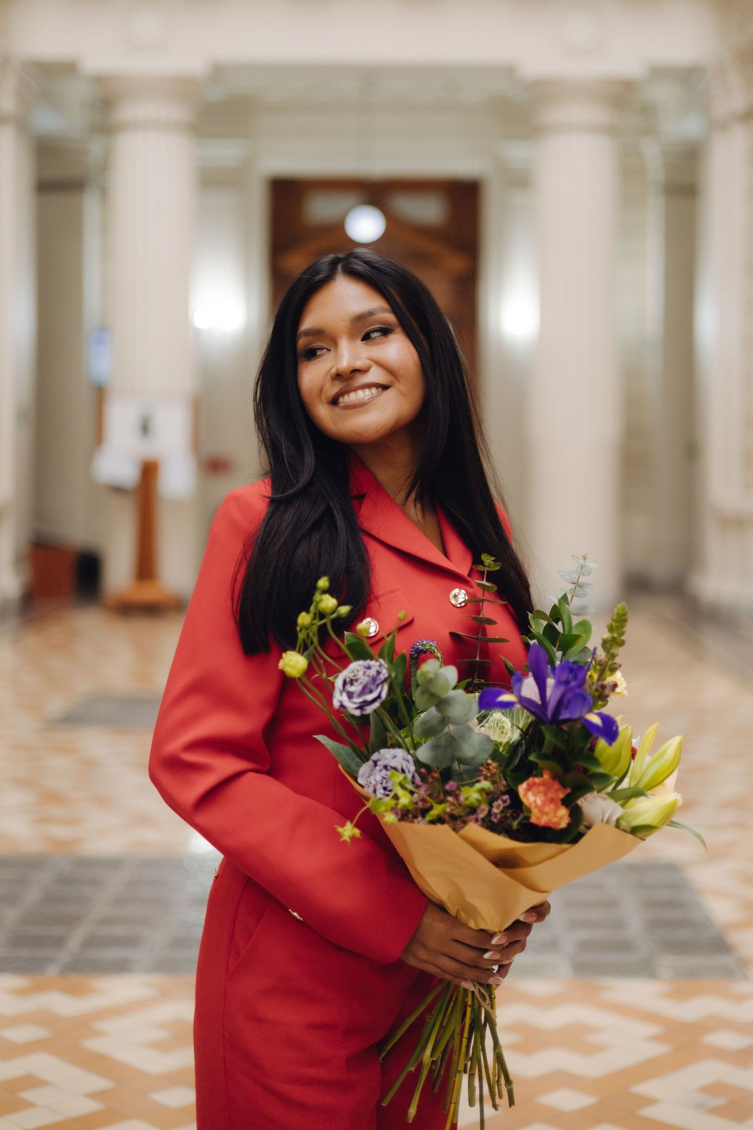 Young woman in red coat smiling while holding vibrant bouquet in ornate hall.
