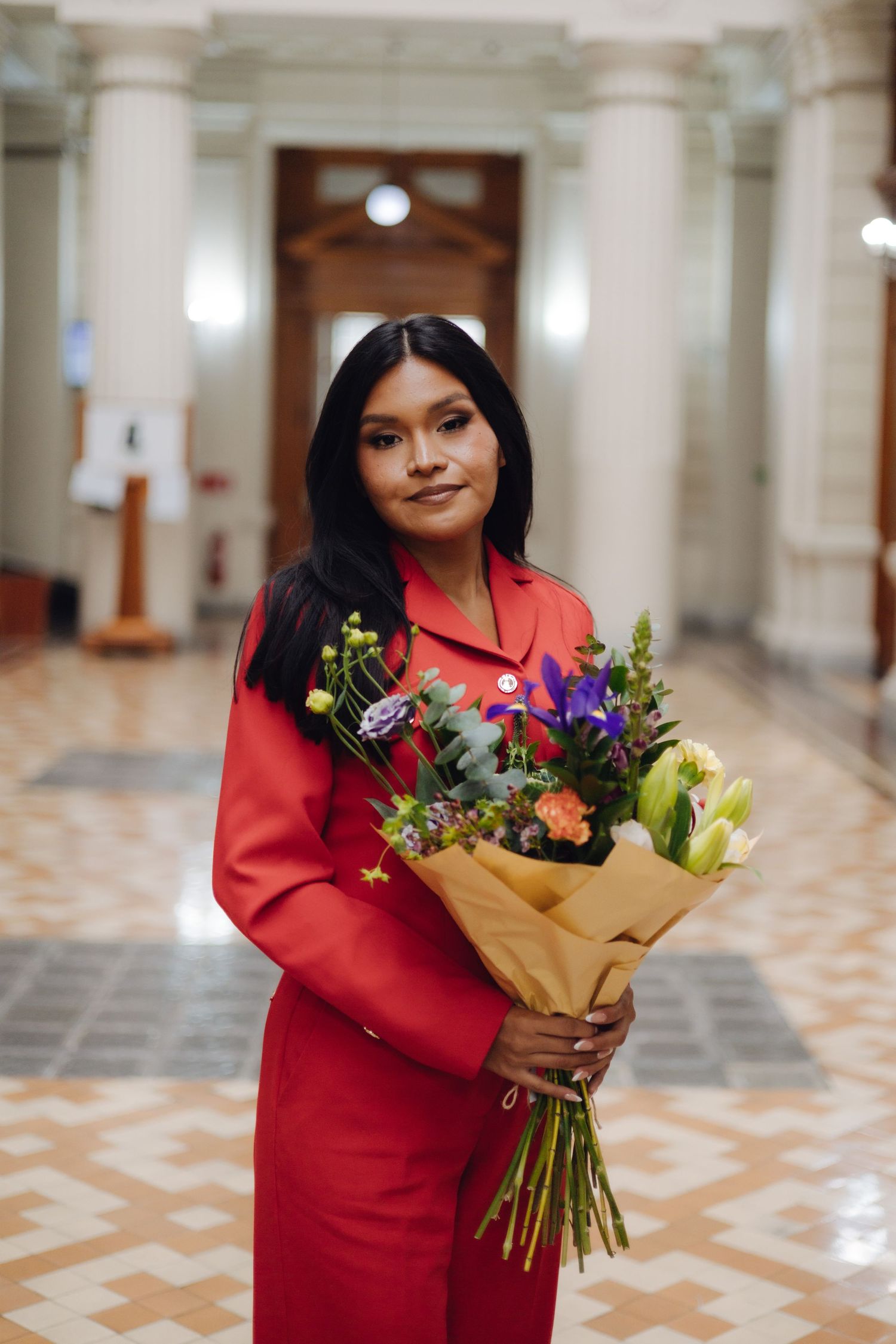 Young woman in red blazer holding mixed flower bouquet in classical building.