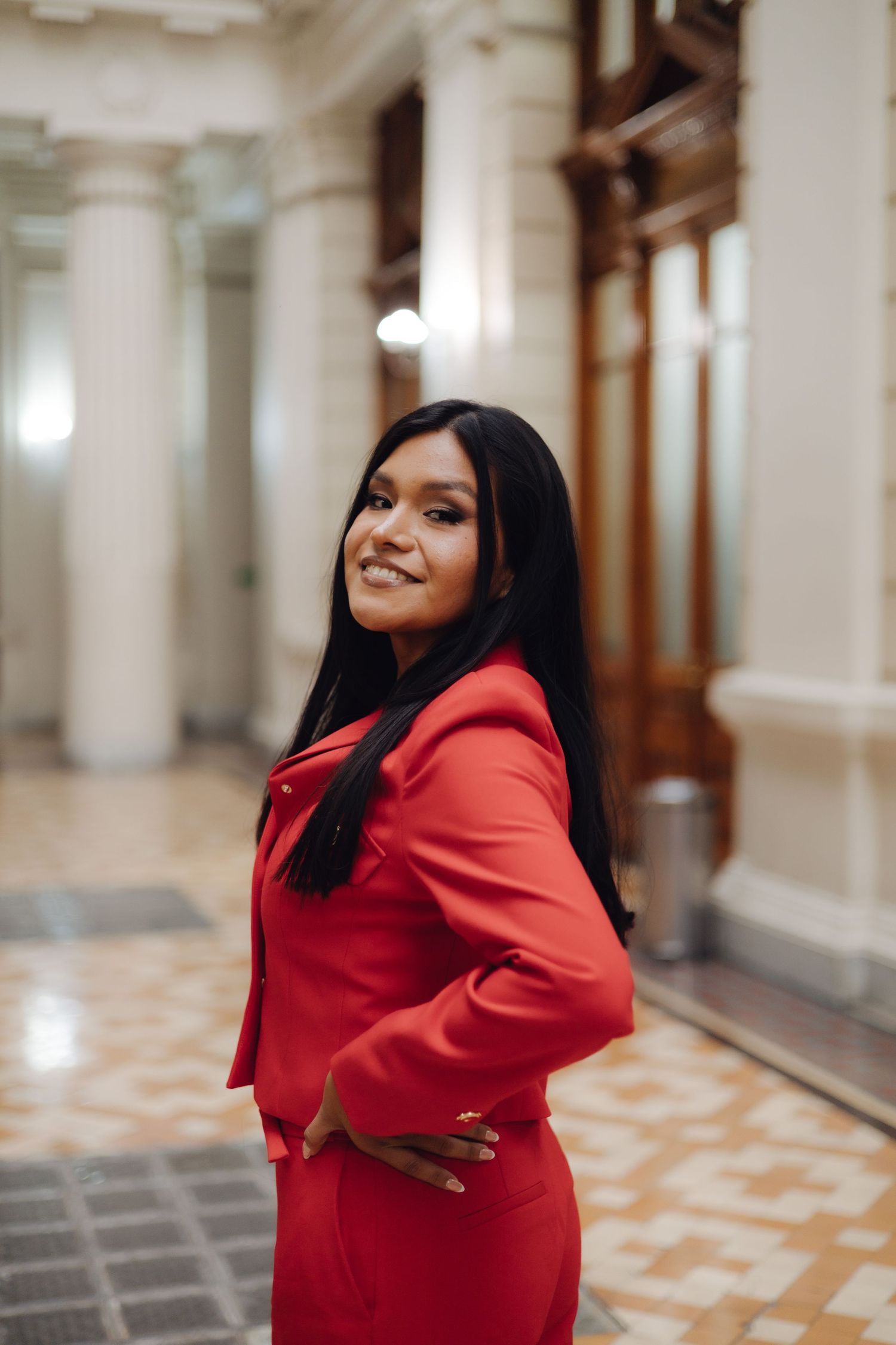 Smiling young woman in red suit posing in elegant columned hallway interior.