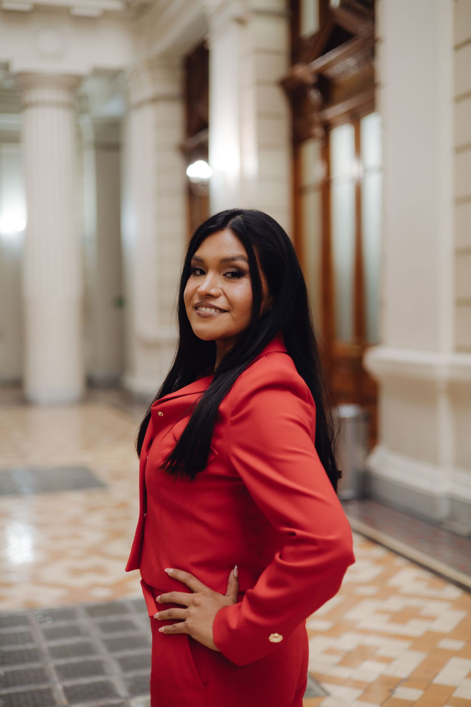 Young woman in red outfit smiling in ornate classical building corridor.