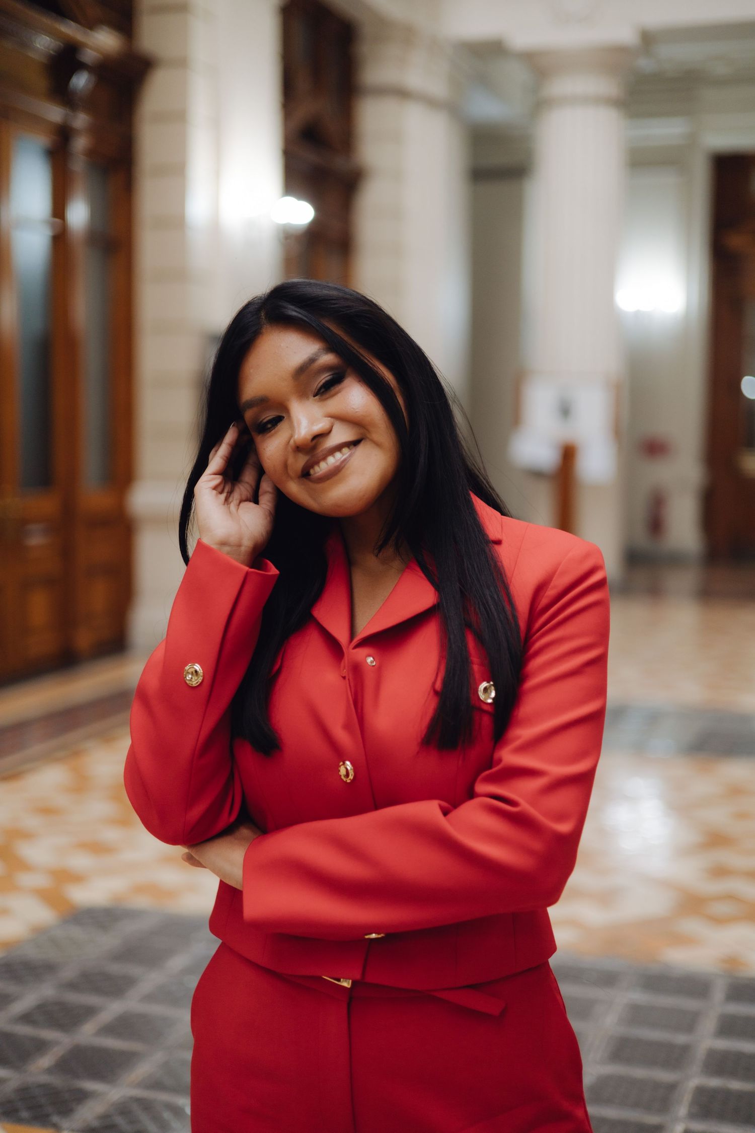 Young woman in red suit smiling with hand near face in elegant hall.