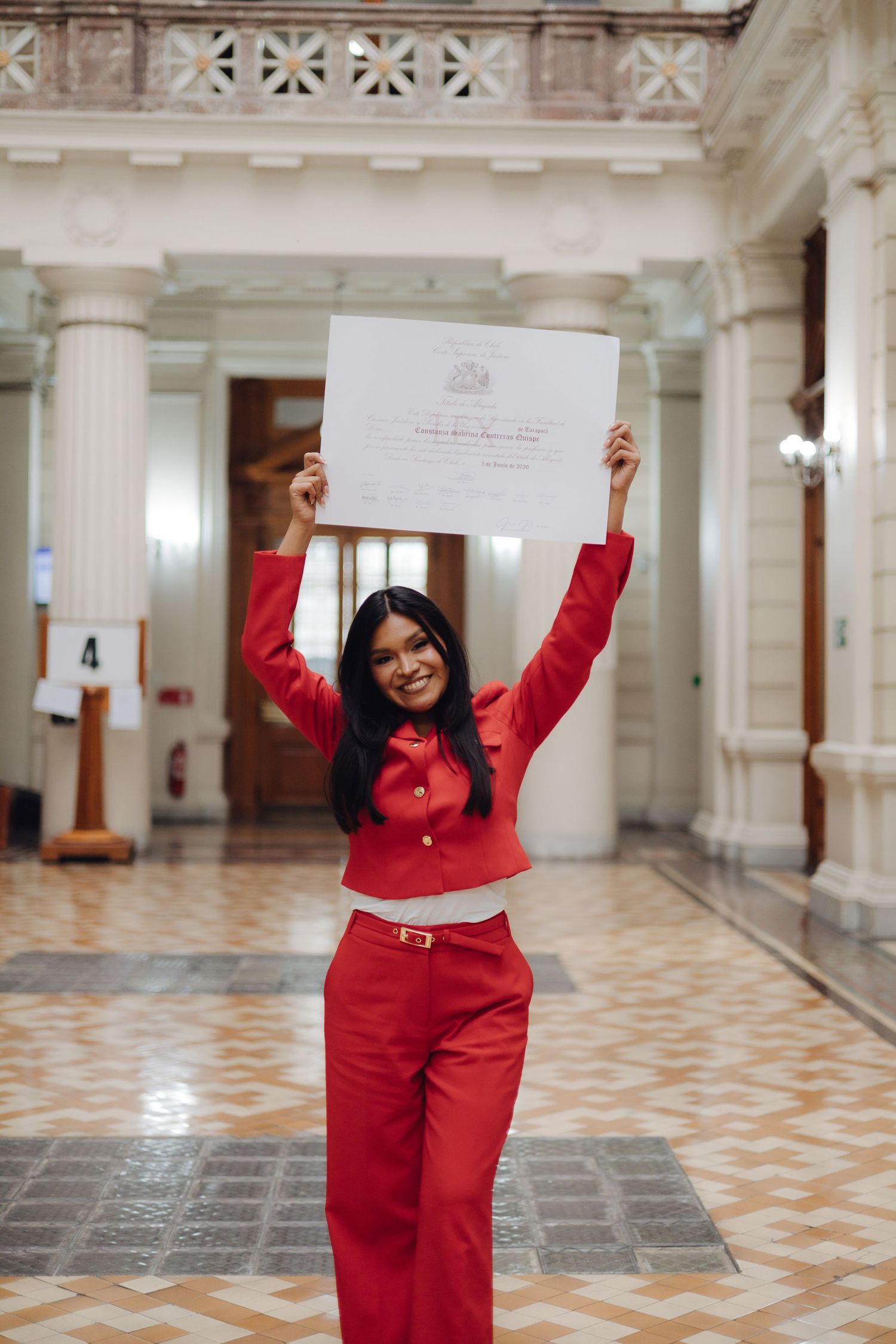 Young woman in red outfit celebrates holding diploma overhead in elegant columned hall.