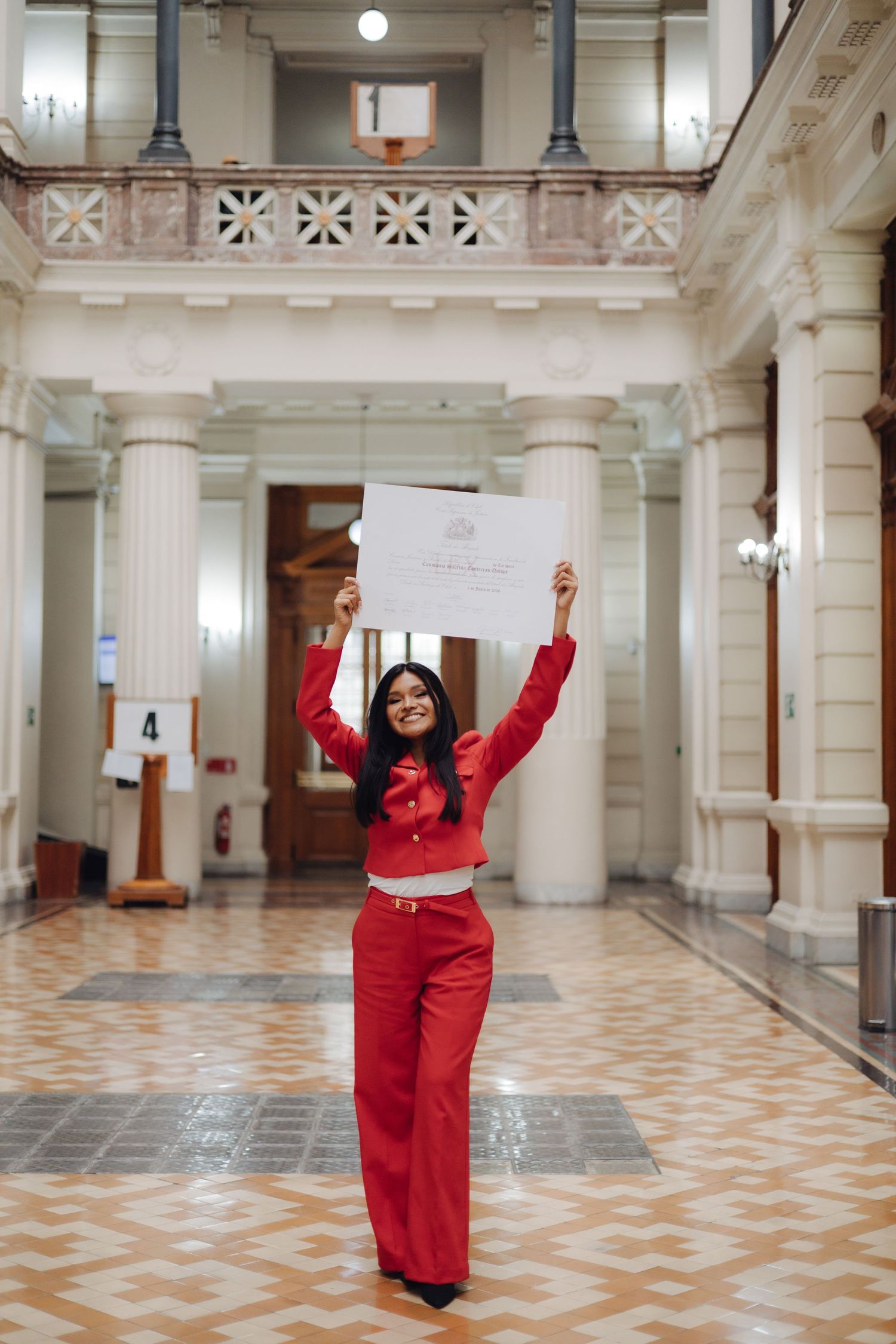 Smiling woman in red suit raises diploma triumphantly in ornate classical building interior.