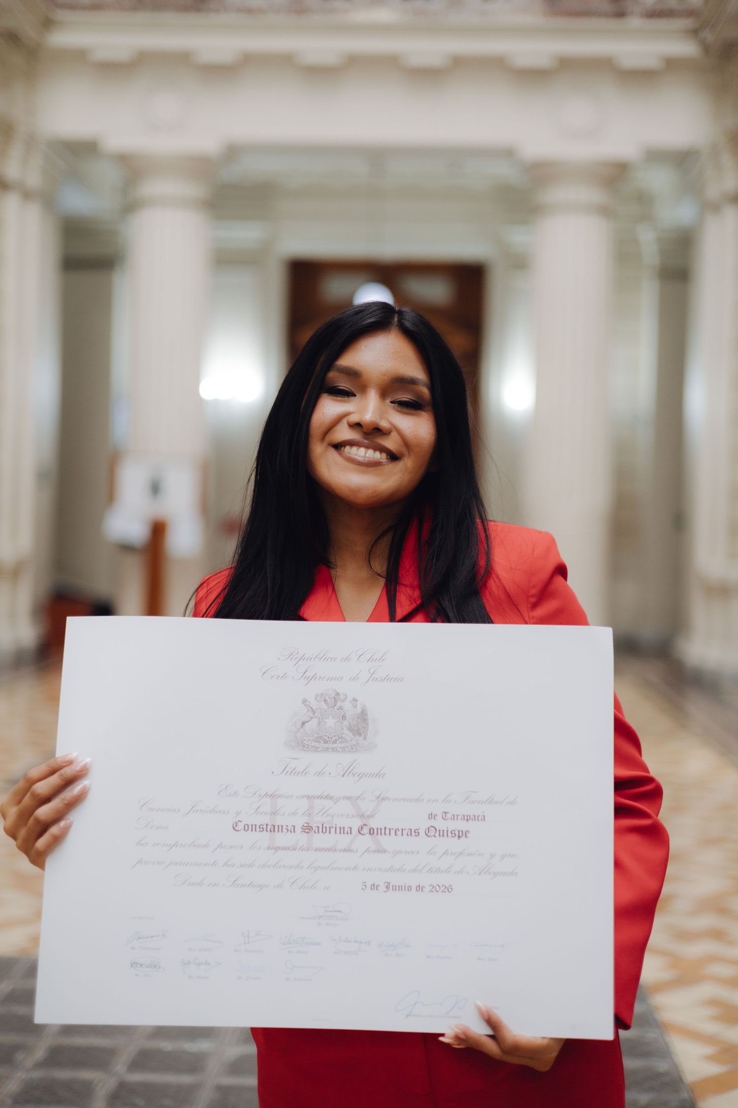 Proud graduate in red blazer displays official diploma certificate in grand columned hall.
