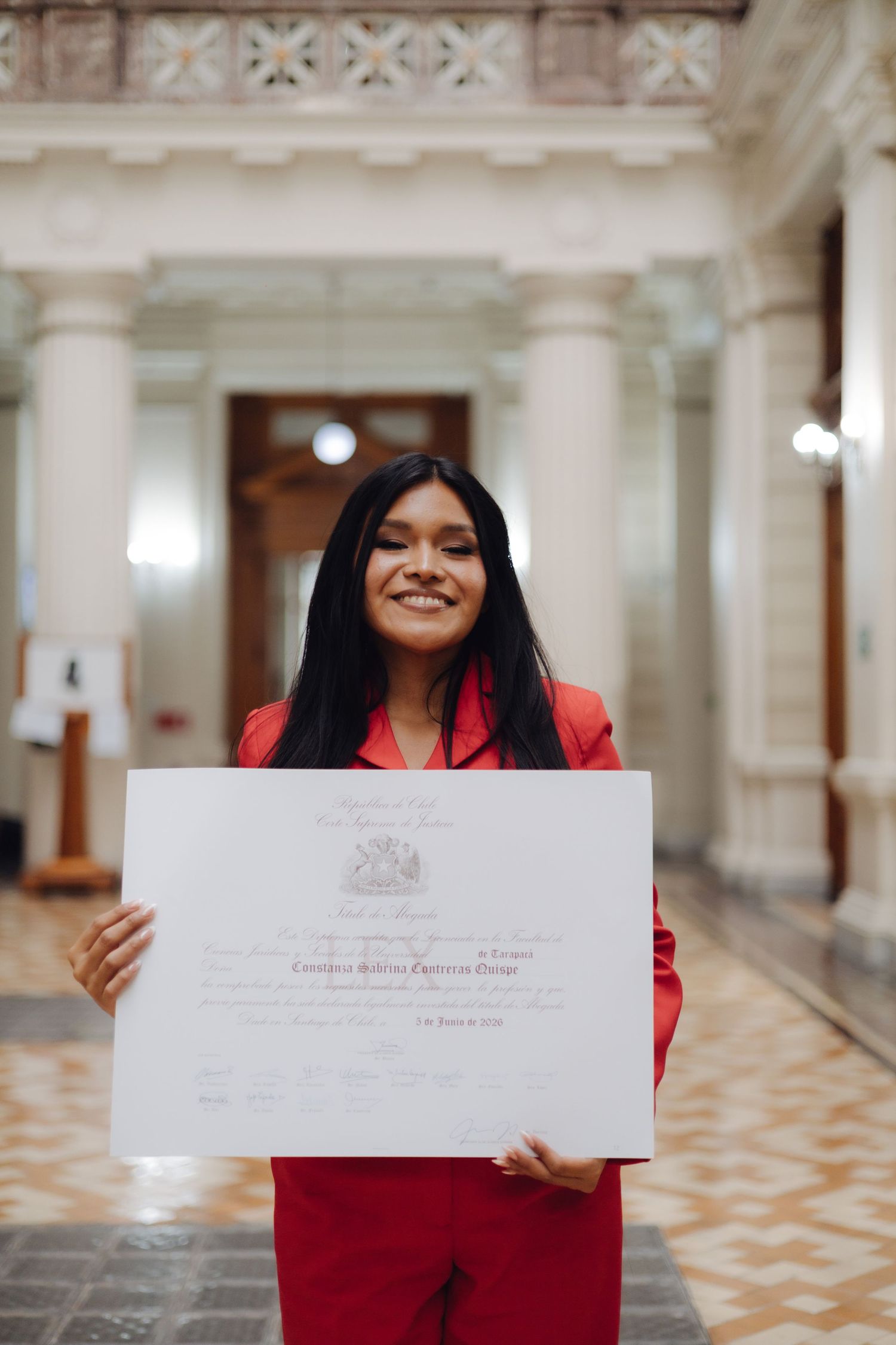Happy young woman in red outfit proudly holds large official diploma in elegant hall.