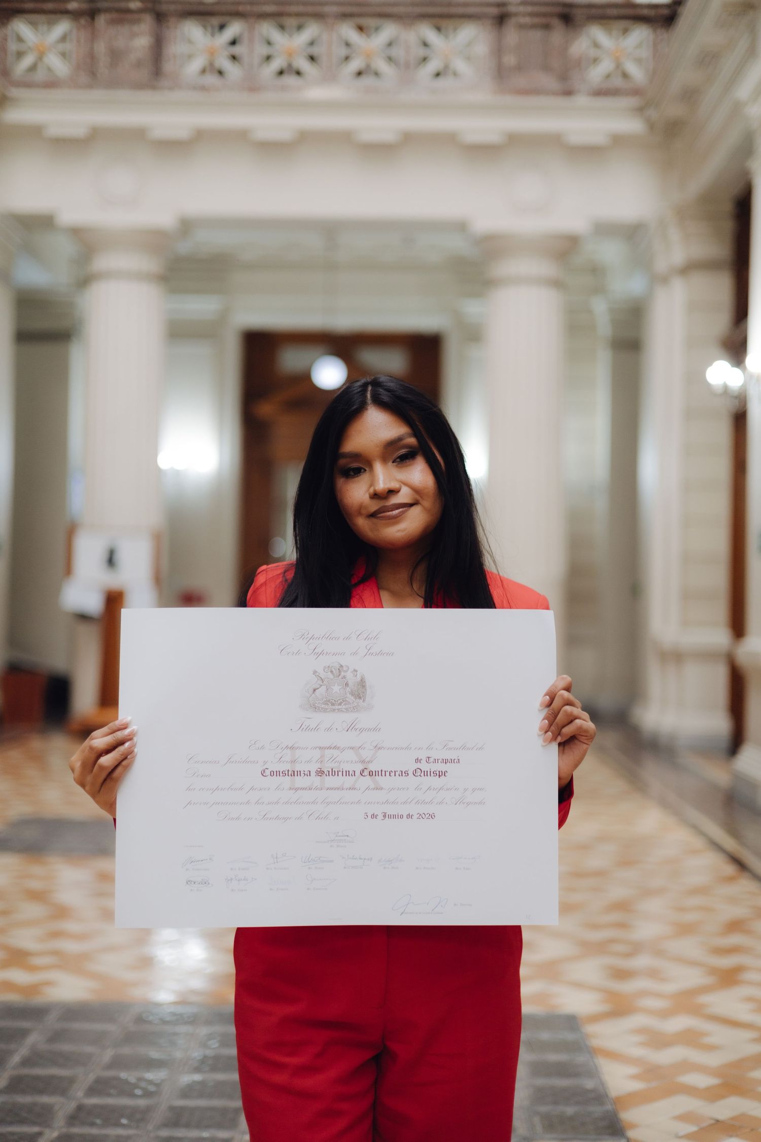 Smiling graduate in red ensemble holds official certificate in beautiful classical building.