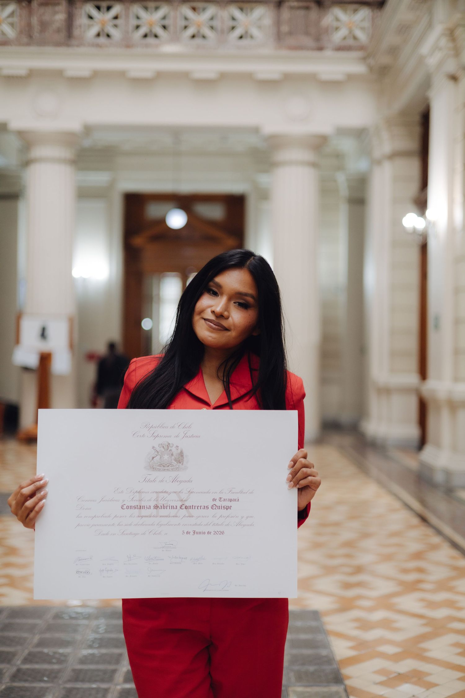 Young woman in red suit holds diploma with subtle smile in grand columned hallway.