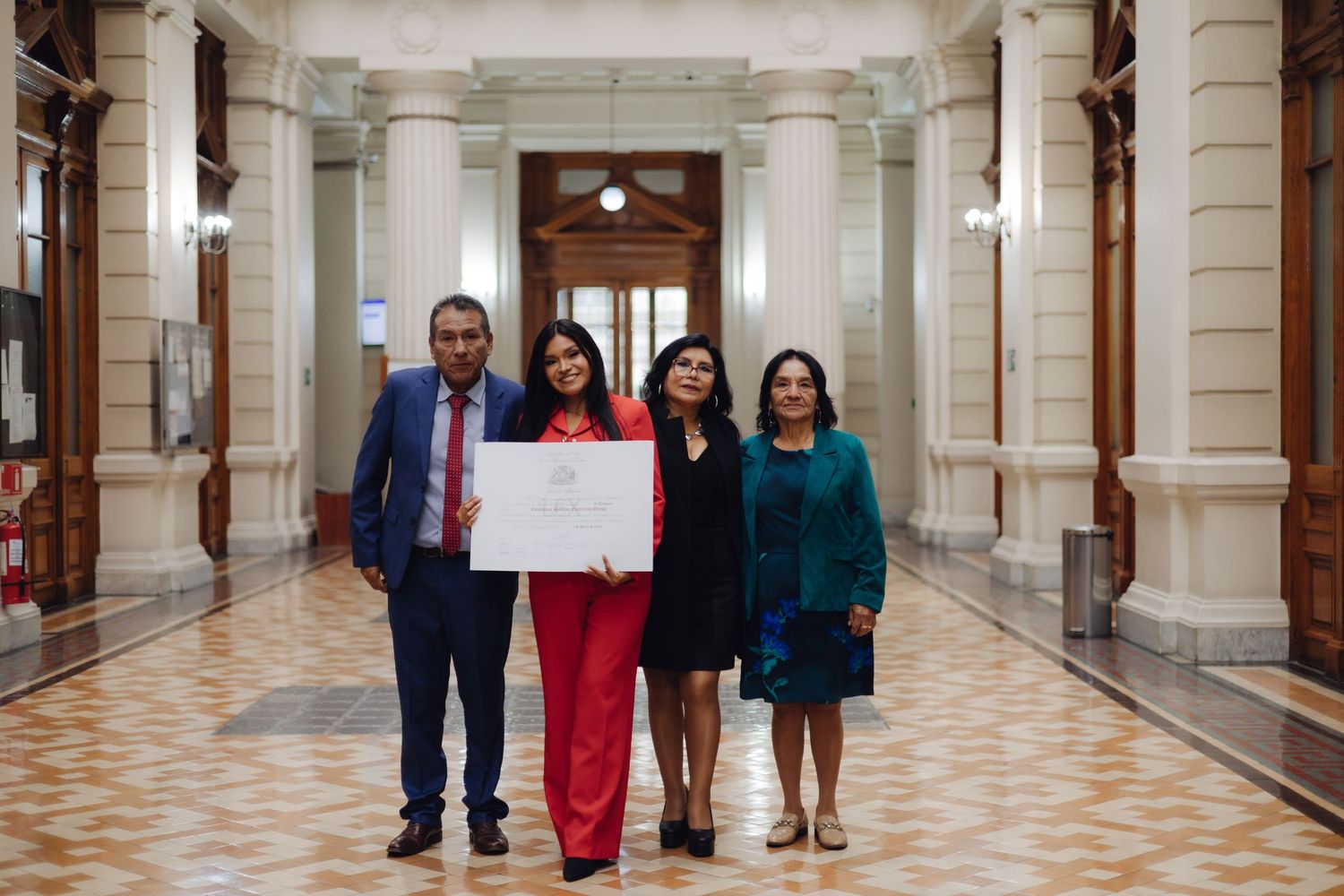 Graduate in red suit poses with three family members holding diploma in ornate corridor.