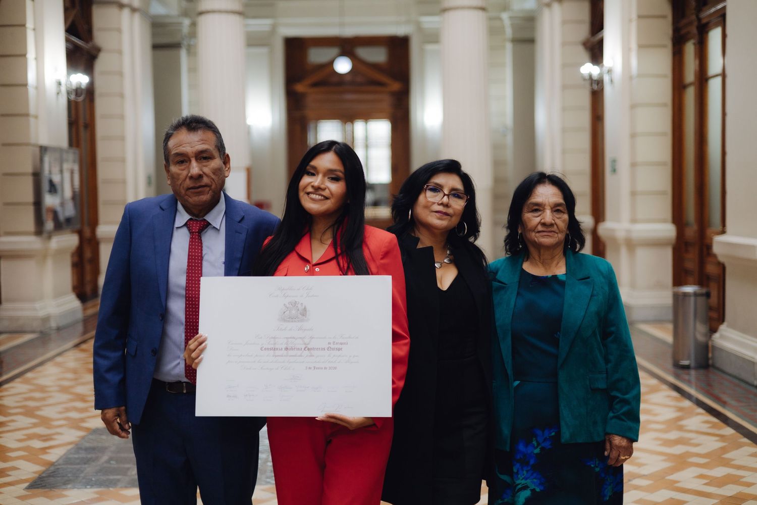 Graduate in red dress poses formally with three family members holding diploma in hall.