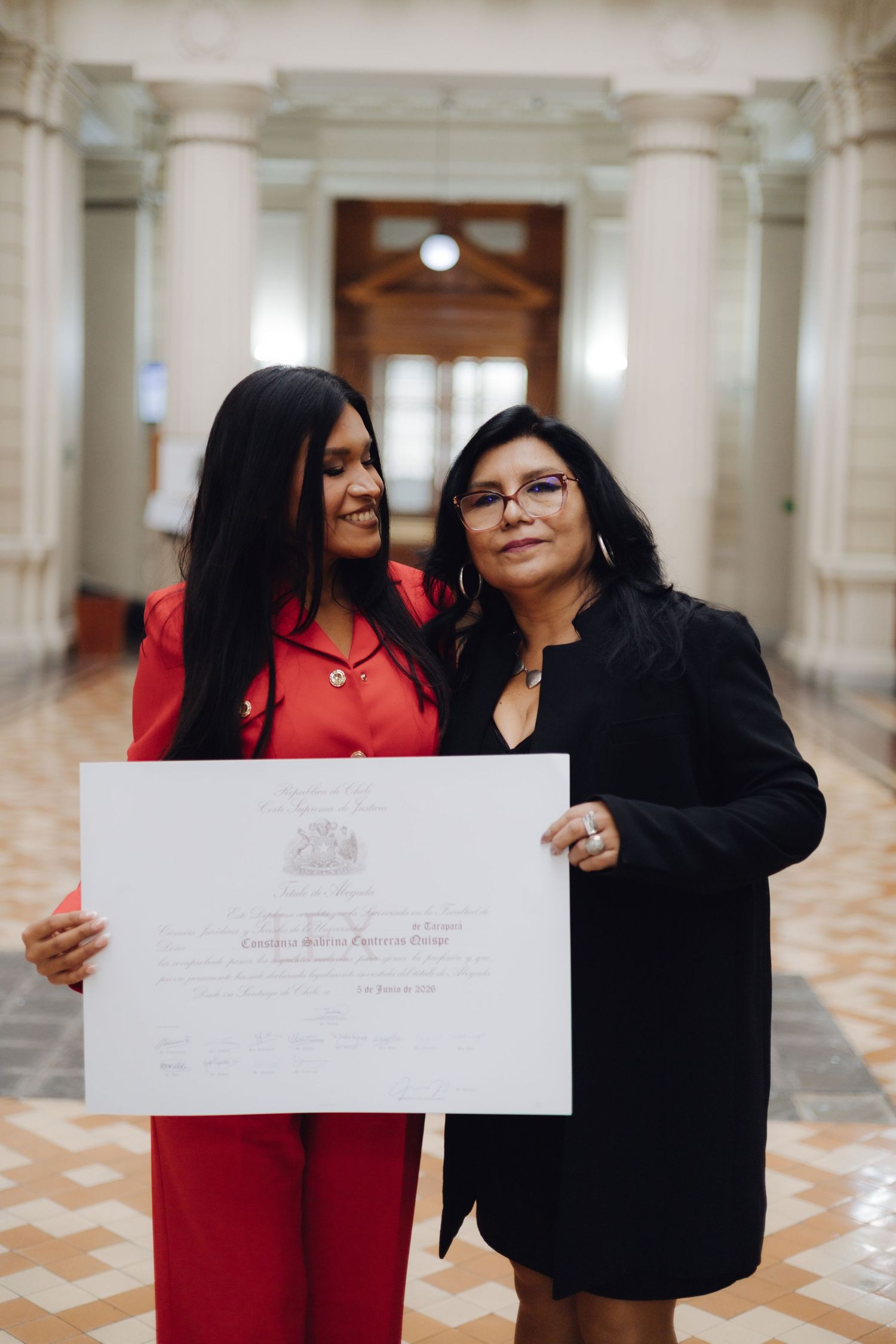 Graduate in red outfit and woman in black share diploma celebration in columned hall.