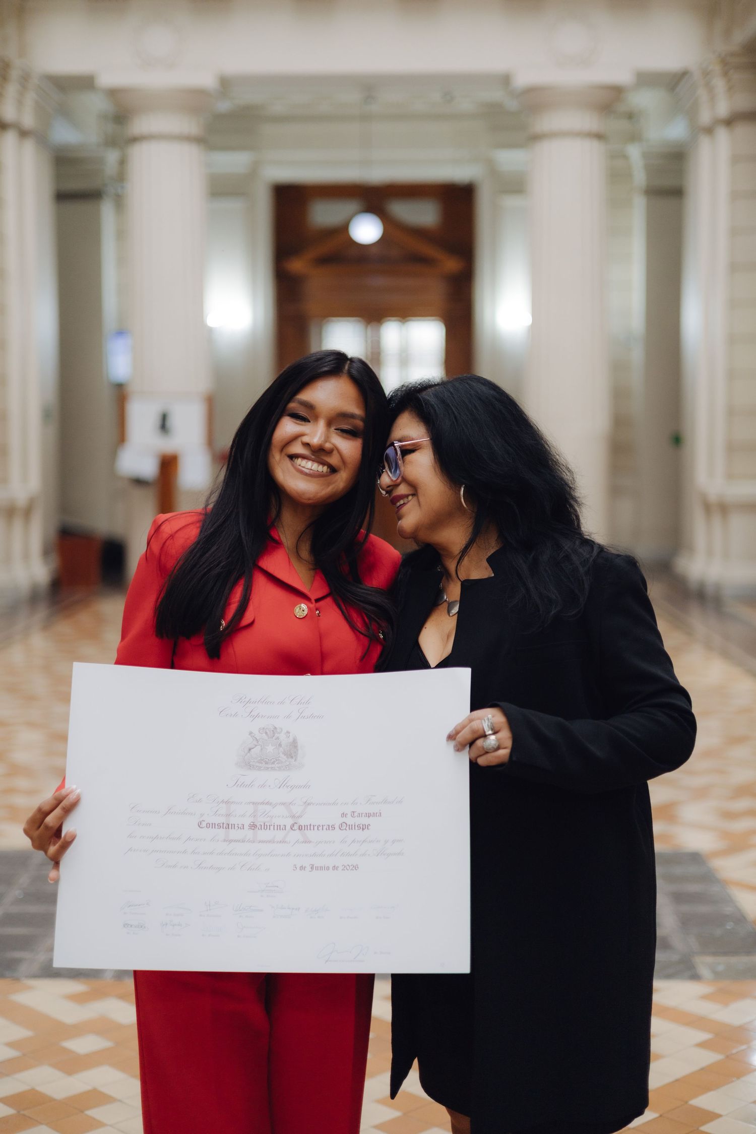 Young woman in red suit and older woman in black jacket proudly holding a diploma certificate in an elegant hallway.