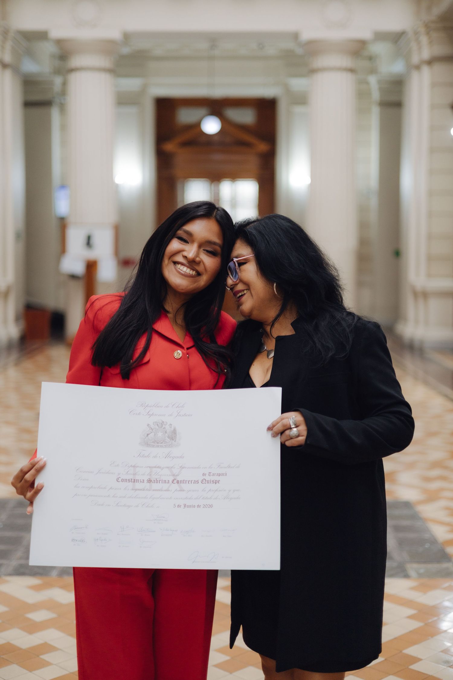 Two women smiling and holding a graduation diploma certificate together in a grand architectural corridor.