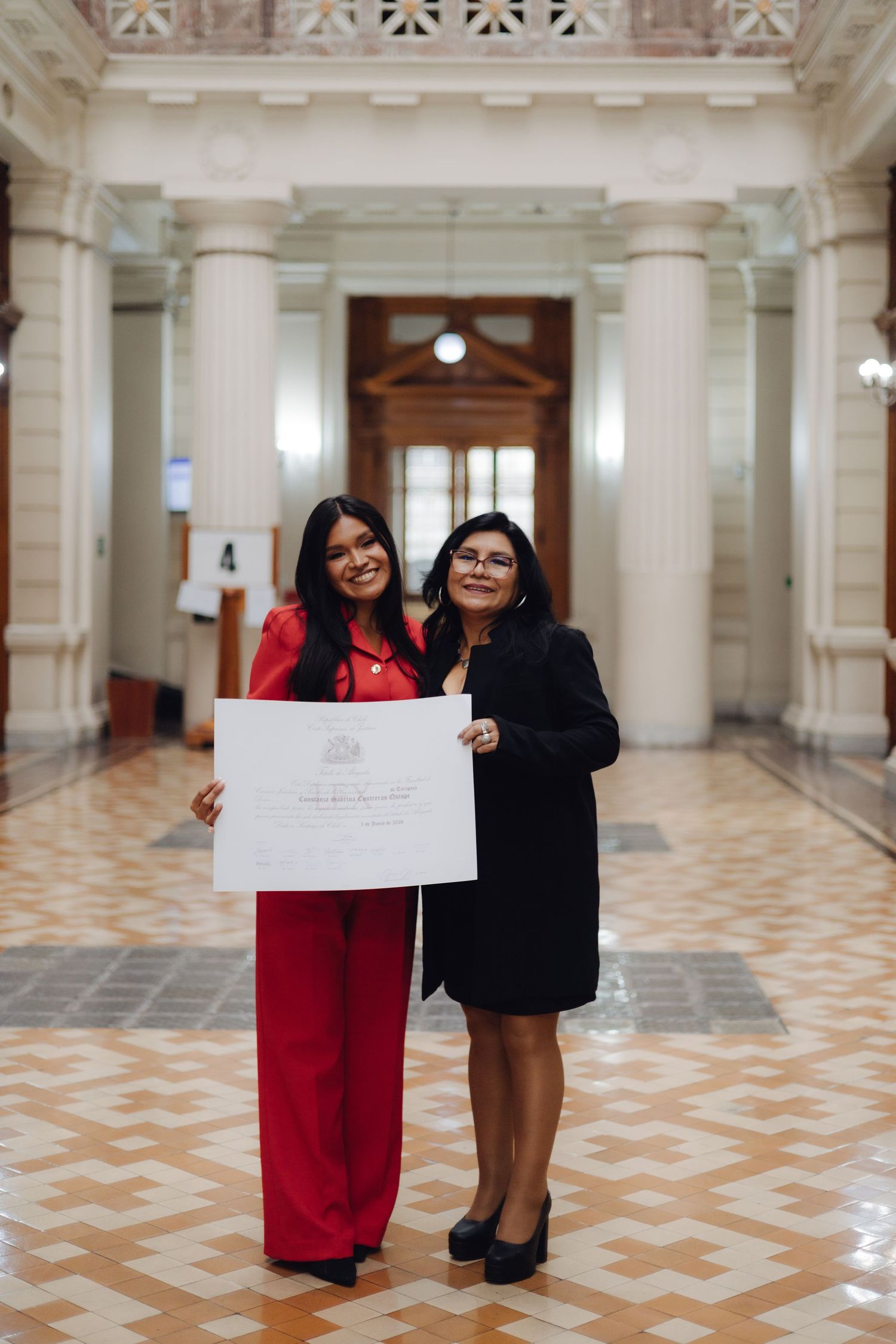 Two women posing with a diploma certificate in a formal building with marble floors and tall columns.