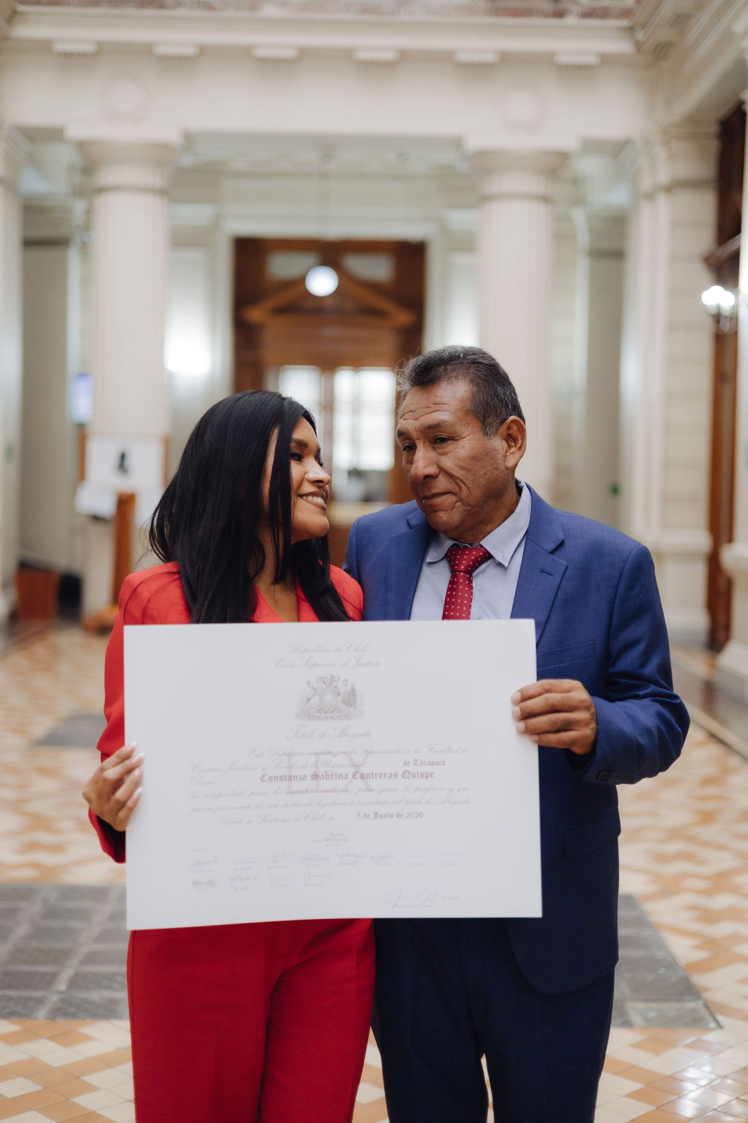 Young woman in red suit and older man in blue suit holding a diploma certificate together indoors.