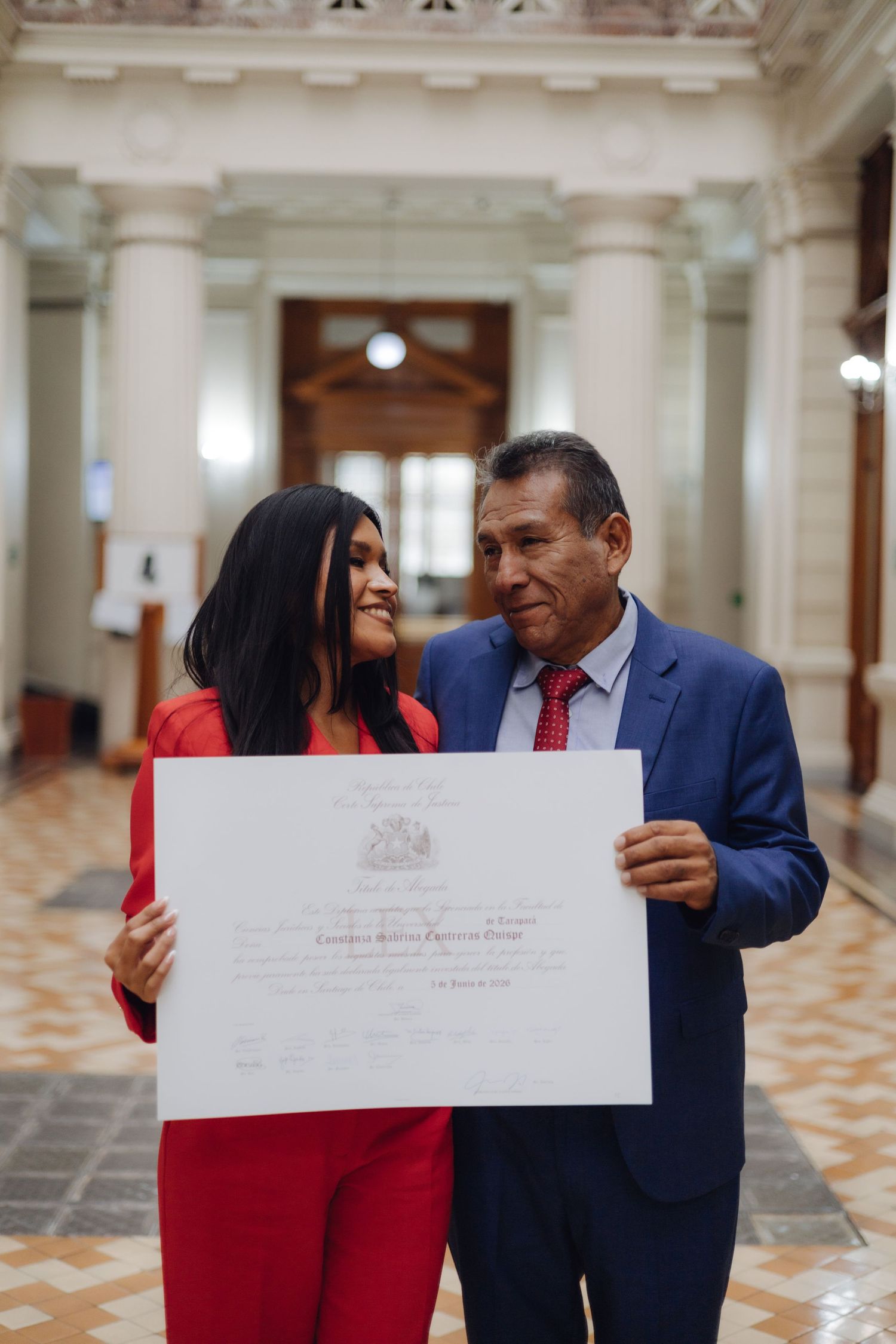 Woman in red outfit and man in blue suit sharing a proud moment holding a graduation diploma certificate.