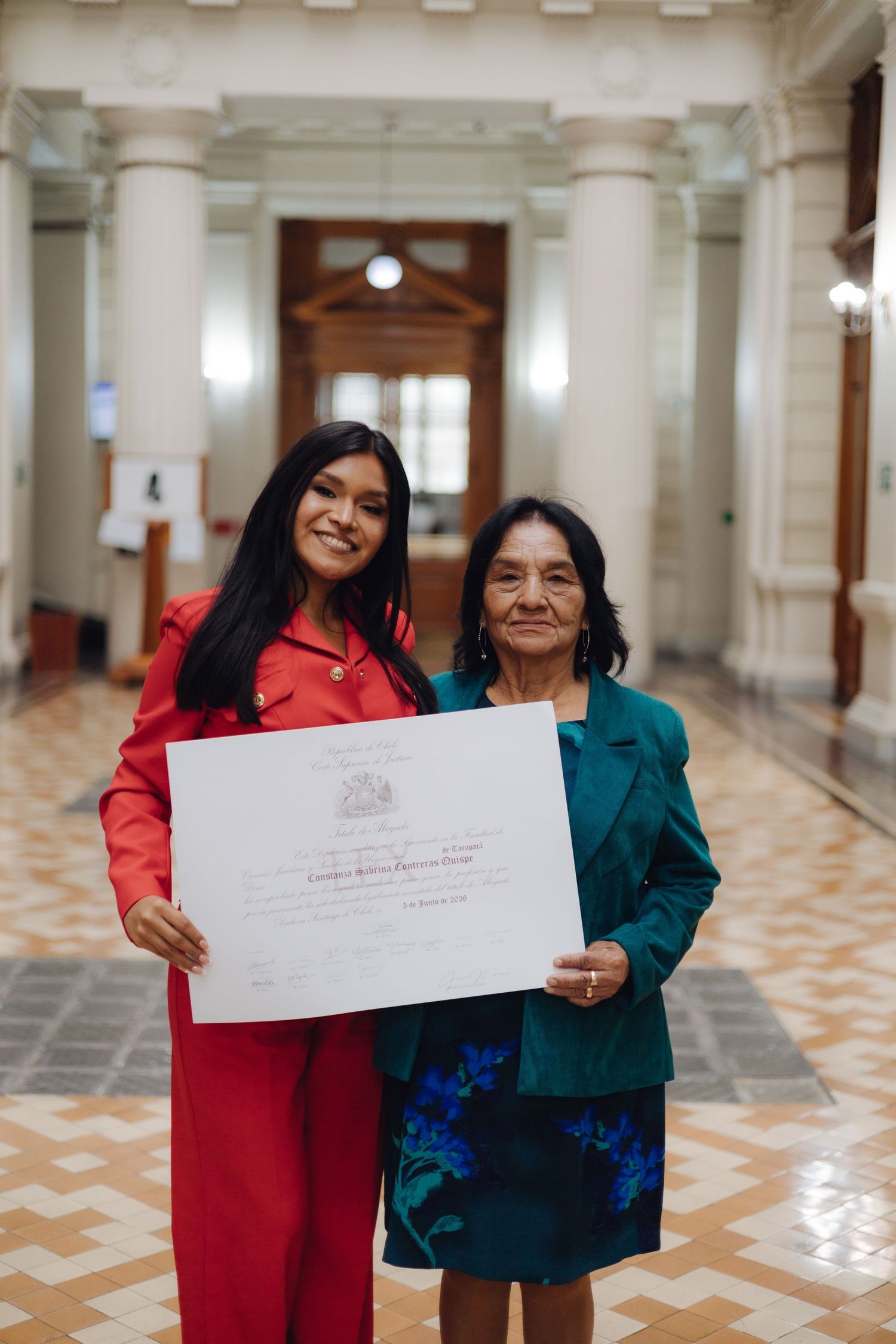 Young graduate in red suit posing with elderly woman in teal jacket holding a diploma certificate.