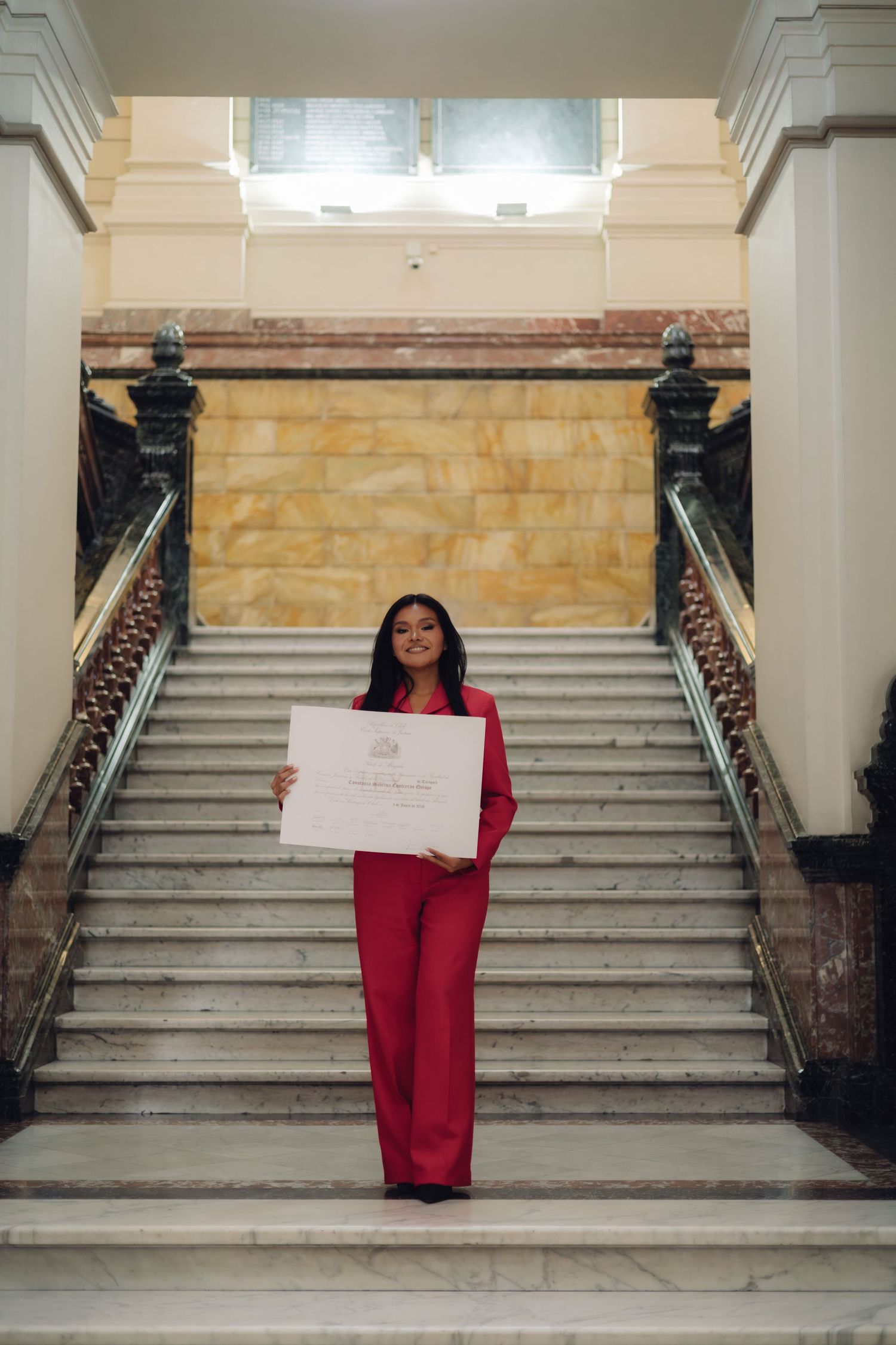 Young woman in red suit standing on grand marble staircase holding a diploma certificate proudly.