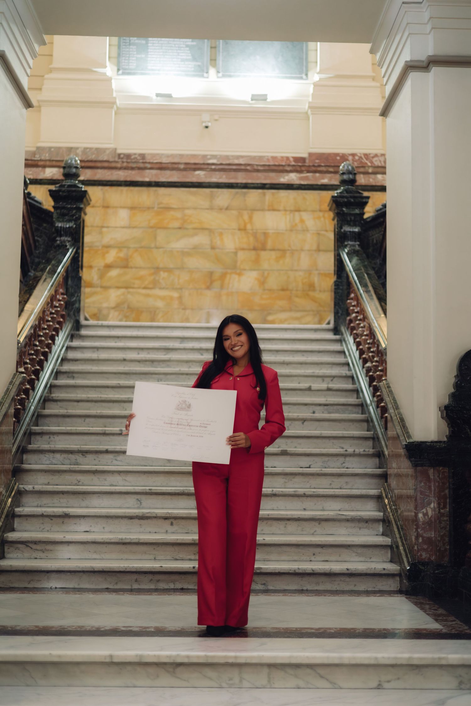 Smiling graduate in red pantsuit holding diploma certificate on elegant marble staircase in formal building.