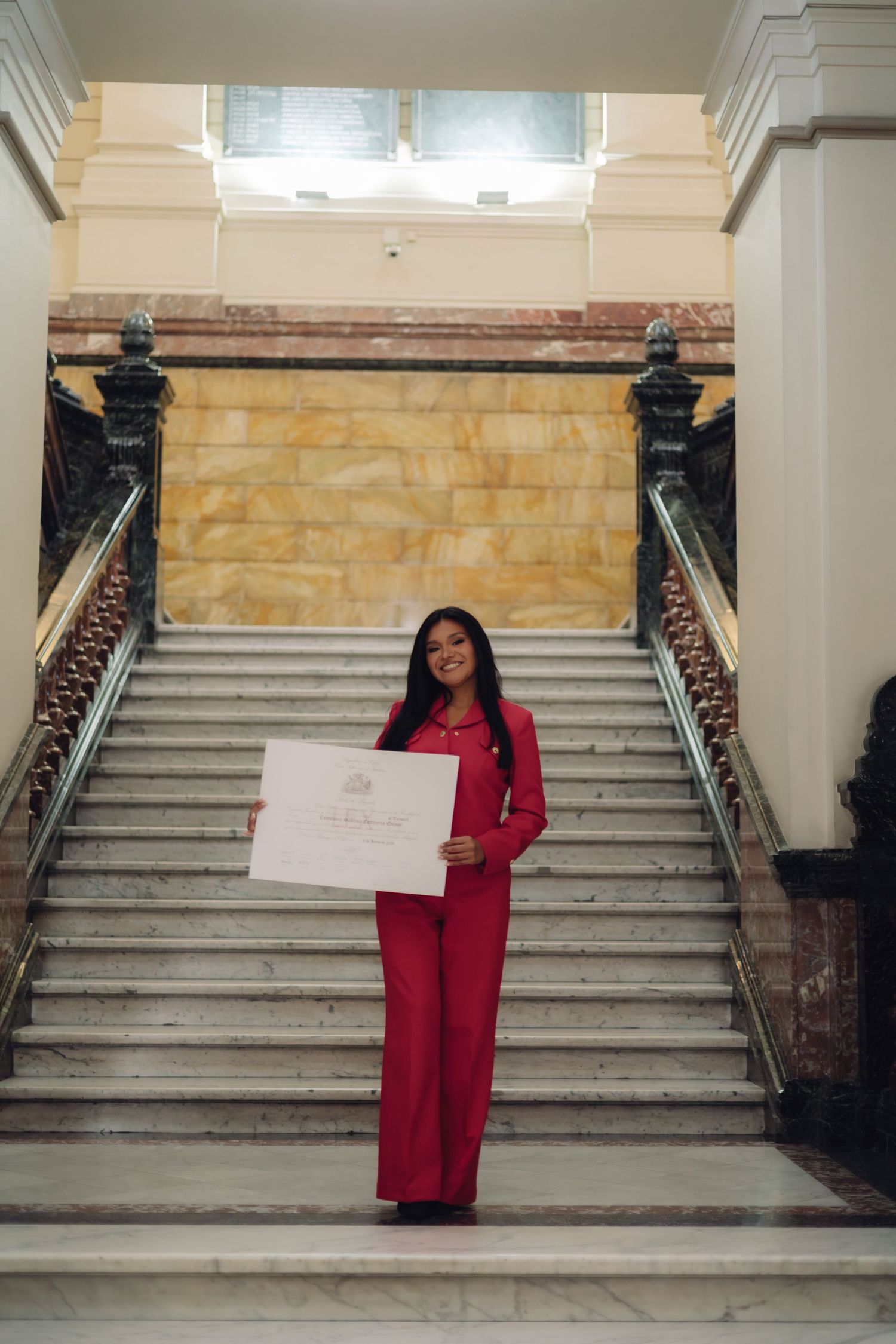 Young woman in red suit proudly displaying a diploma certificate while standing on a grand staircase.