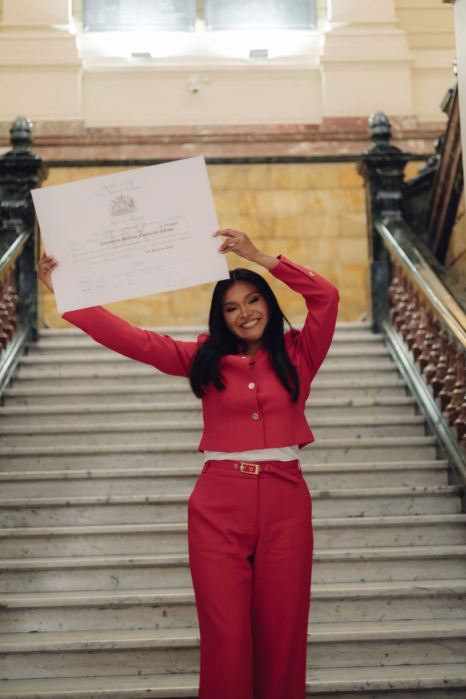 Joyful graduate in red outfit raising diploma certificate overhead on a grand marble staircase.
