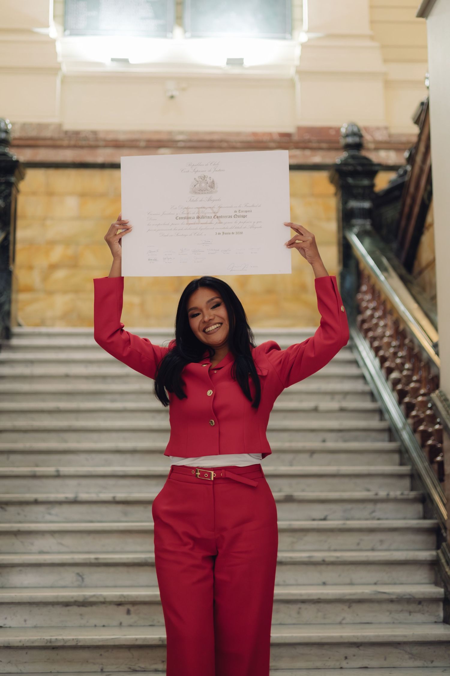 Woman in red suit raises diploma overhead on grand staircase.
