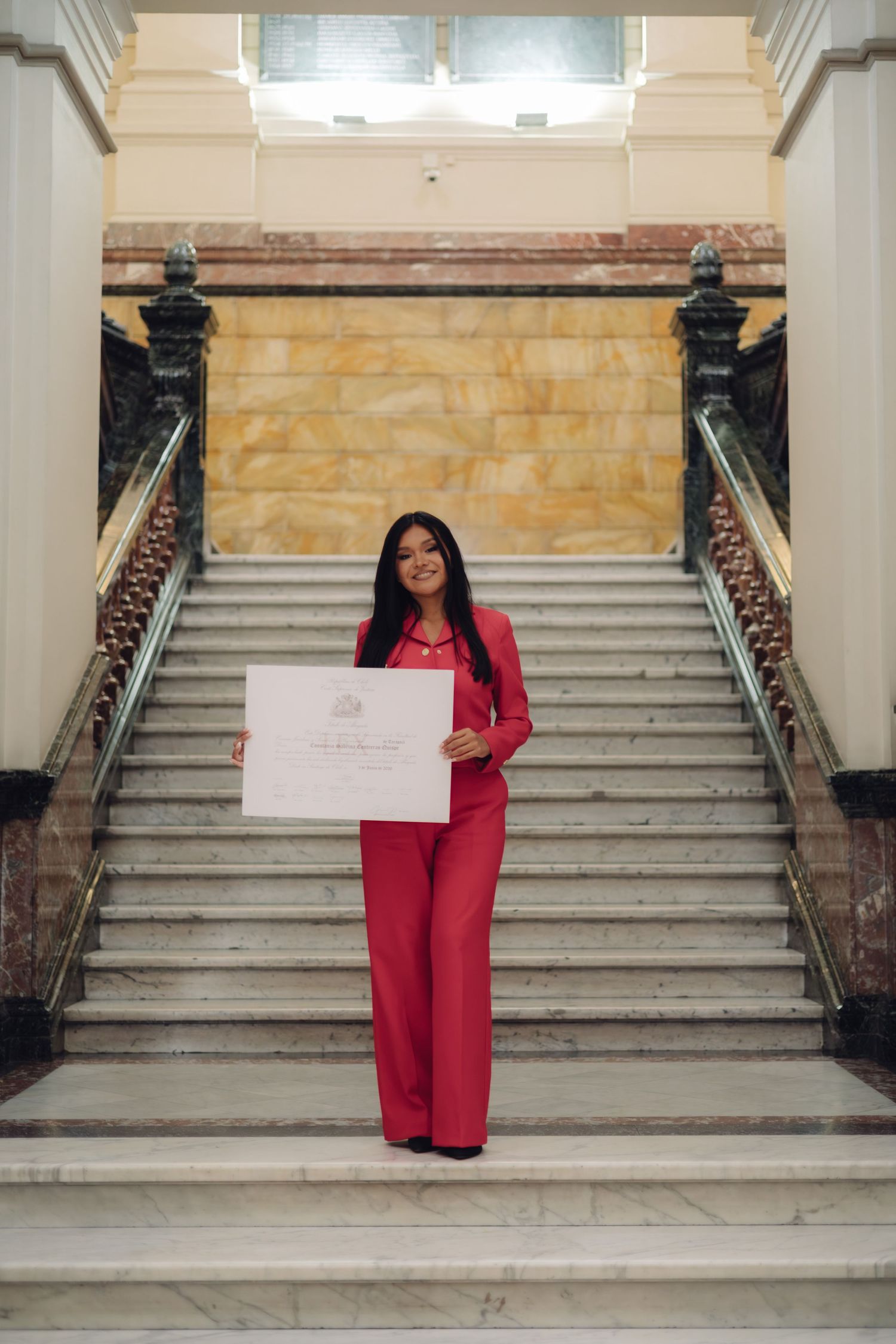 Smiling woman in red pantsuit holds certificate on marble staircase.