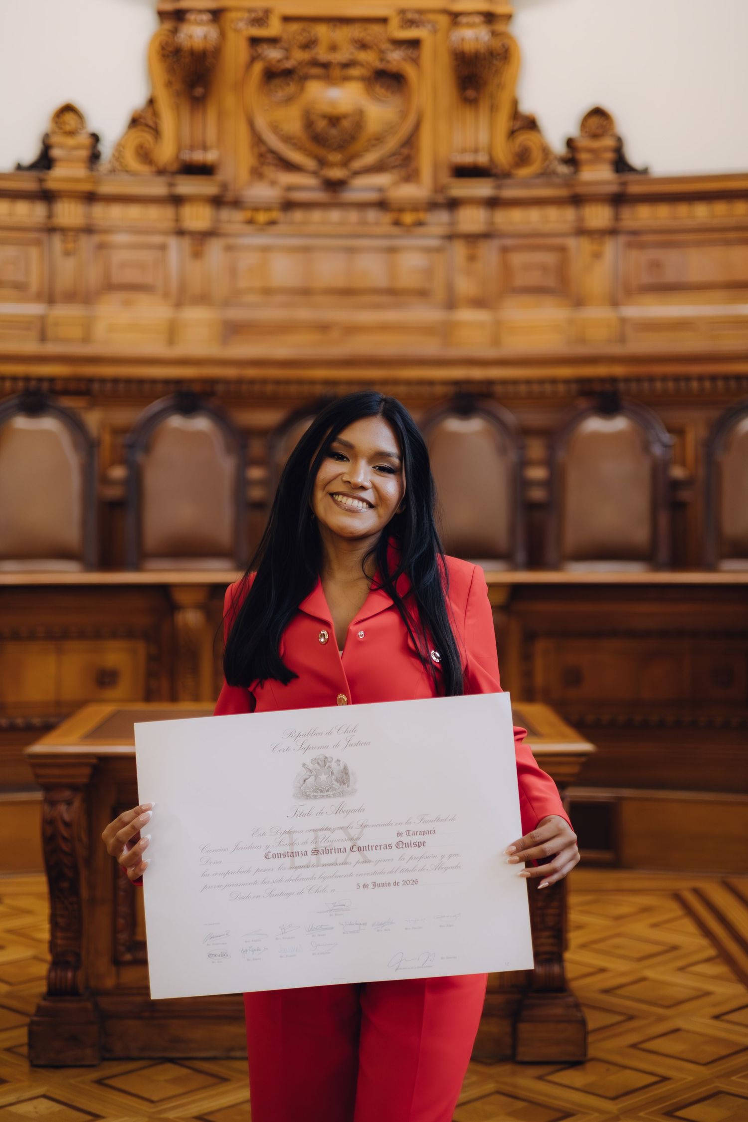 Smiling woman in red suit displays official certificate indoors.