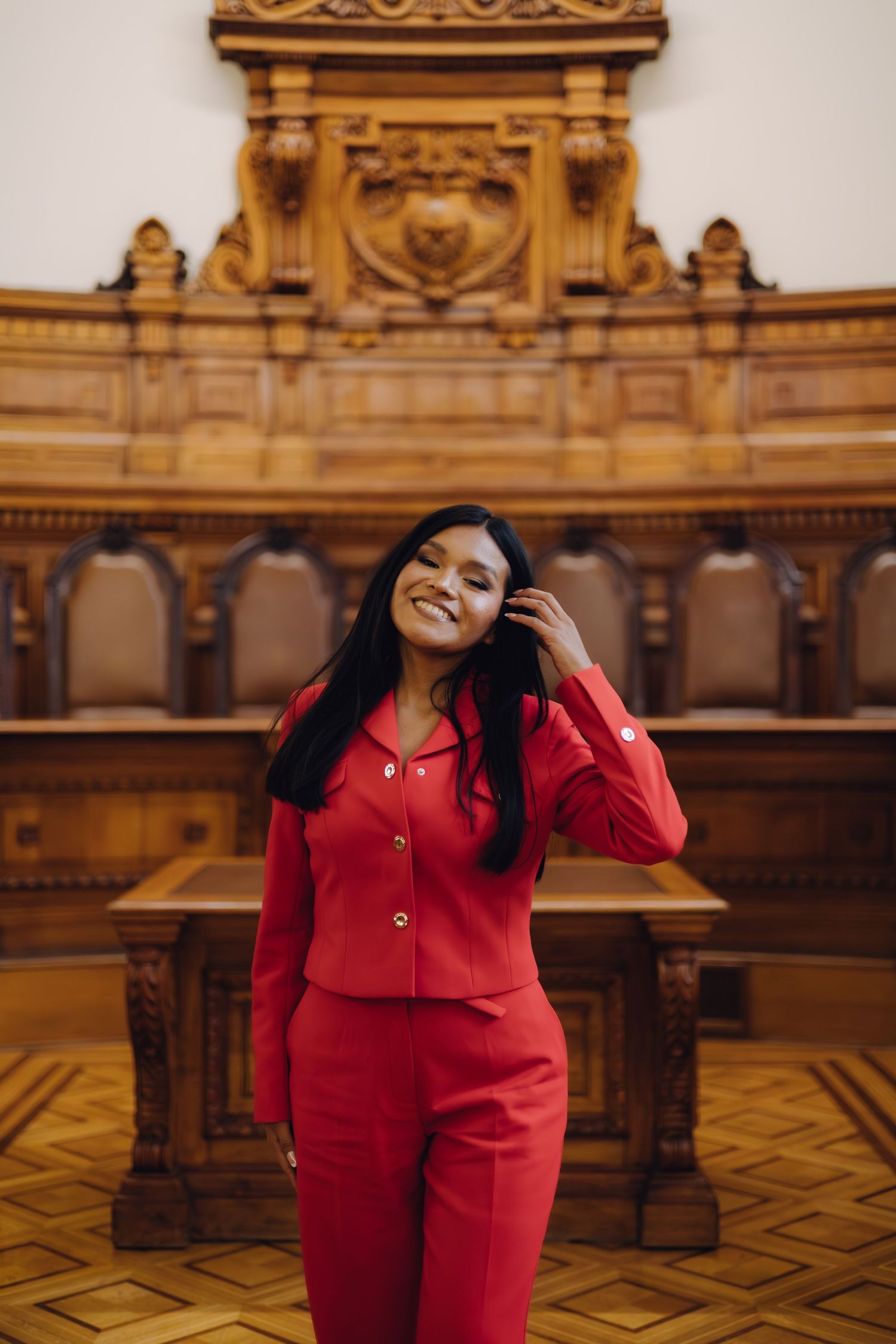 Smiling woman in red suit poses in ornate wood-paneled room.