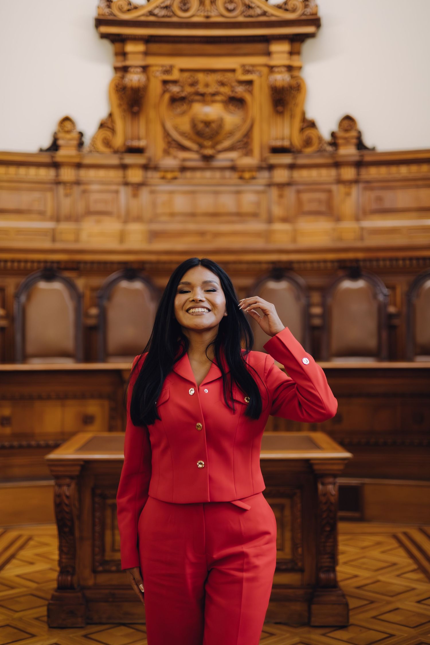 Smiling woman in red suit poses in ornate wooden courtroom interior.