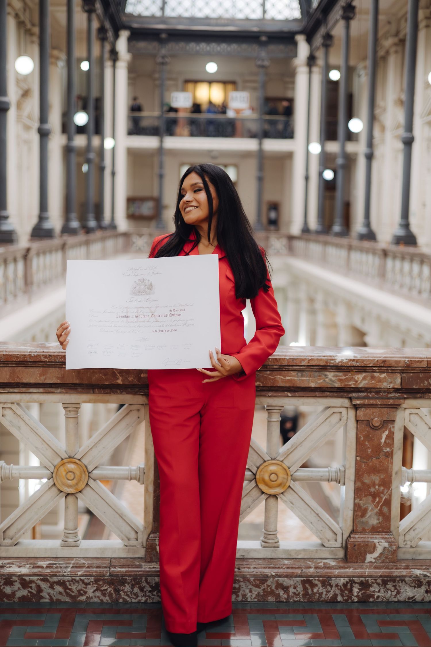 Woman in red jumpsuit holds diploma certificate in elegant marble hall.