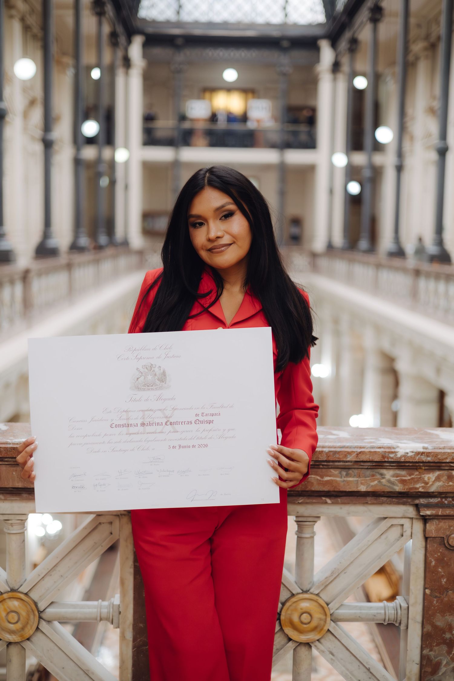 Young woman in red suit proudly displays her graduation diploma indoors.