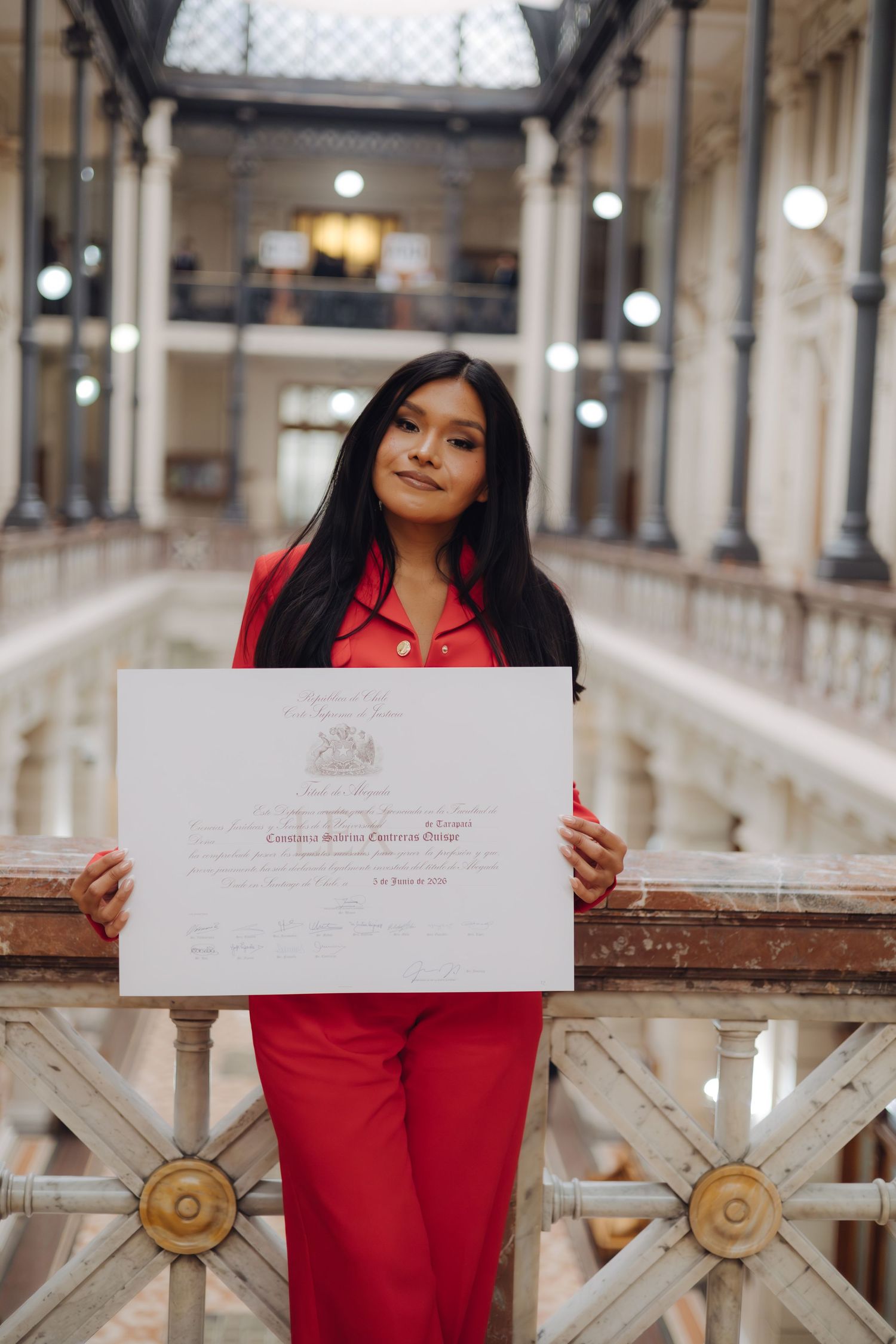 Graduate in red outfit holds official certificate in classic building.