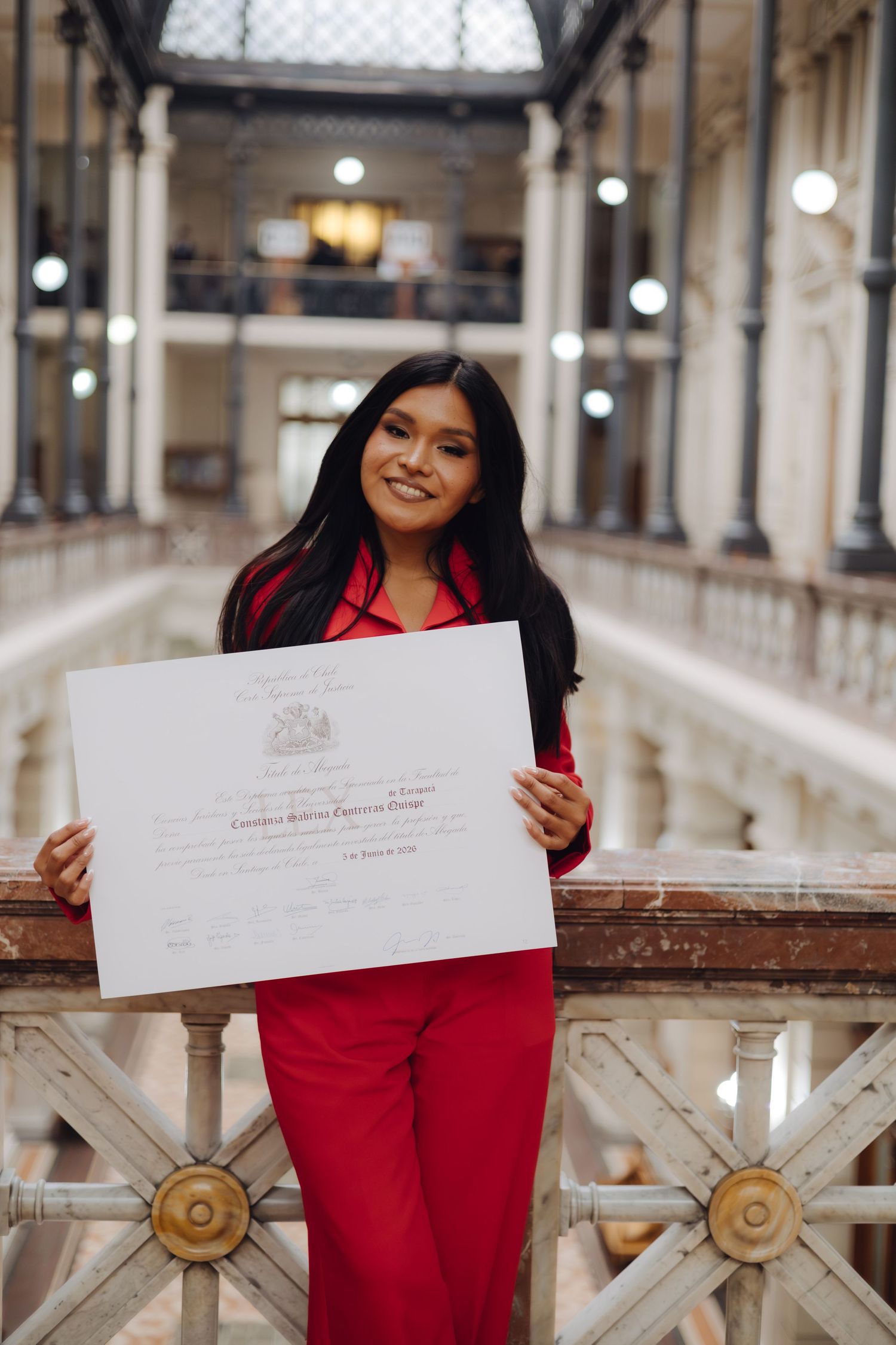 Smiling woman in red suit showcases diploma in grand architectural hall.