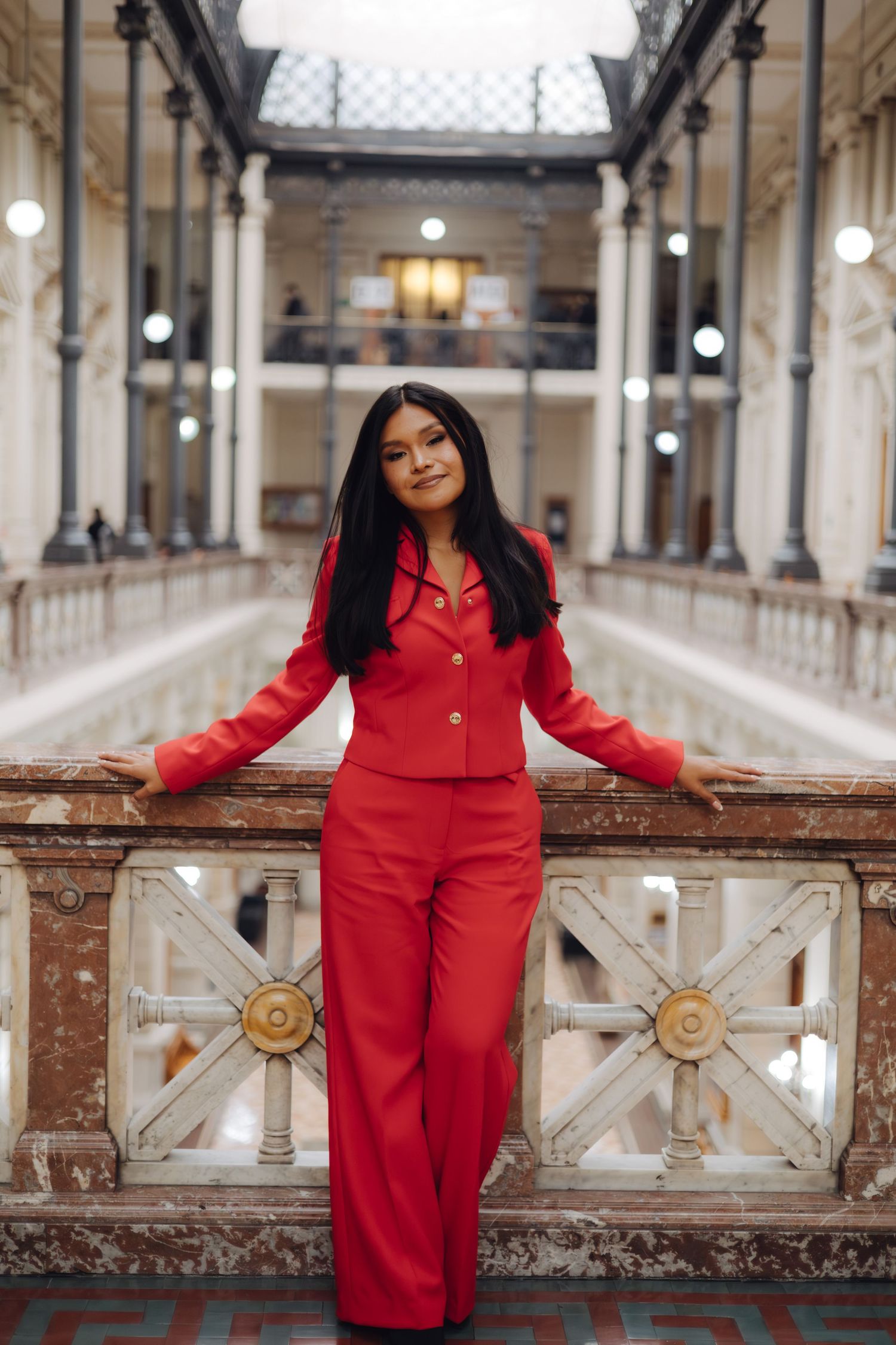 Woman in red suit leans against marble railing in grand gallery.