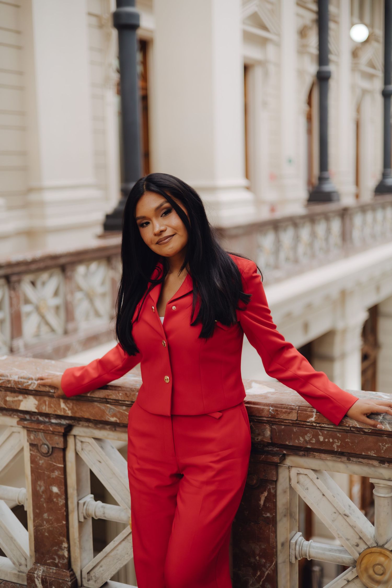 Elegant woman in red blazer and pants poses on ornate balcony.