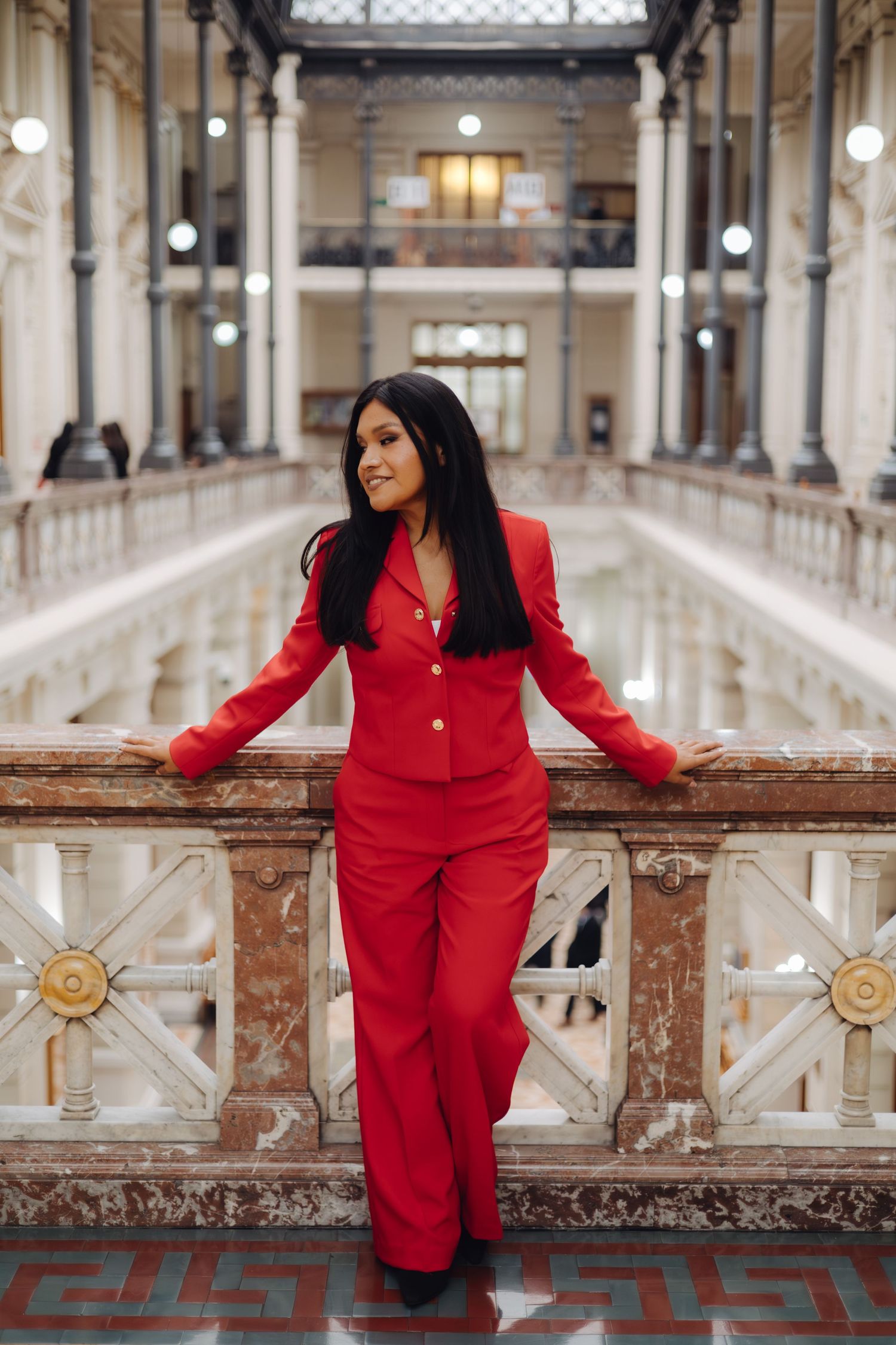 Stylish woman in red outfit poses in elegant marble corridor.