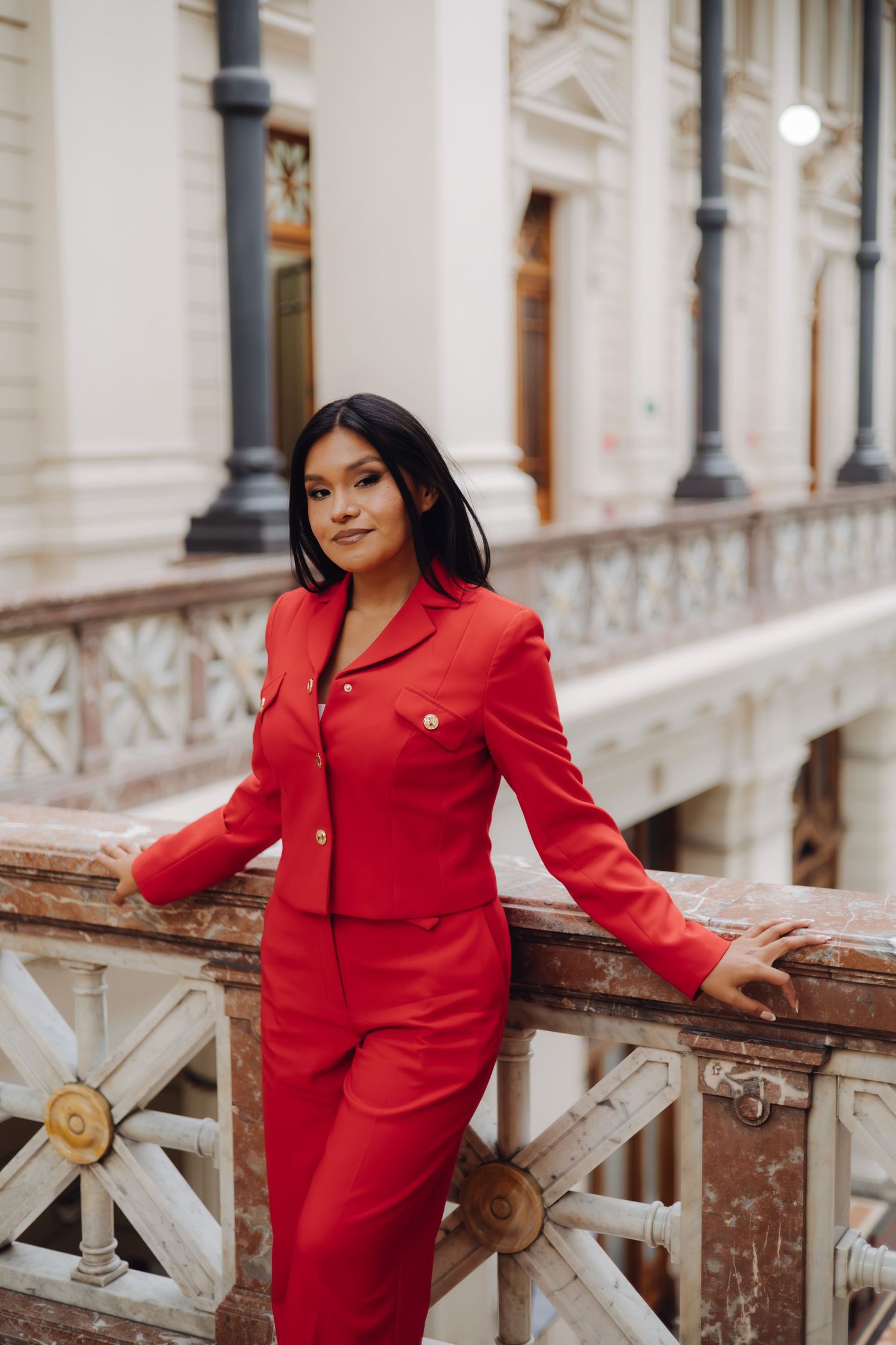 Woman in red blazer and skirt poses on classic building balcony.