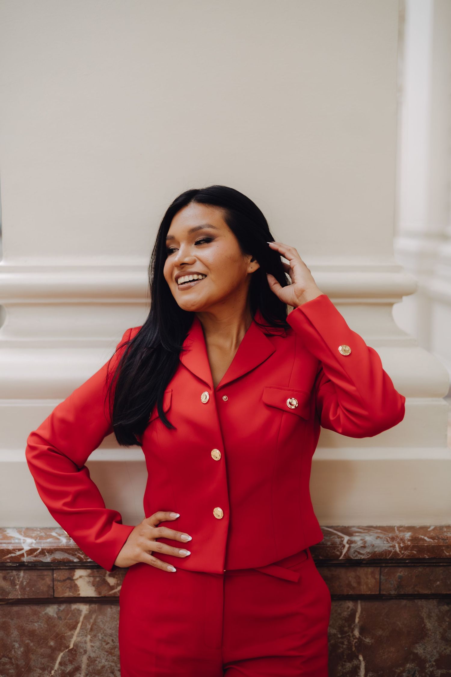Smiling woman in a red blazer suit with gold buttons, posing confidently against a light wall.