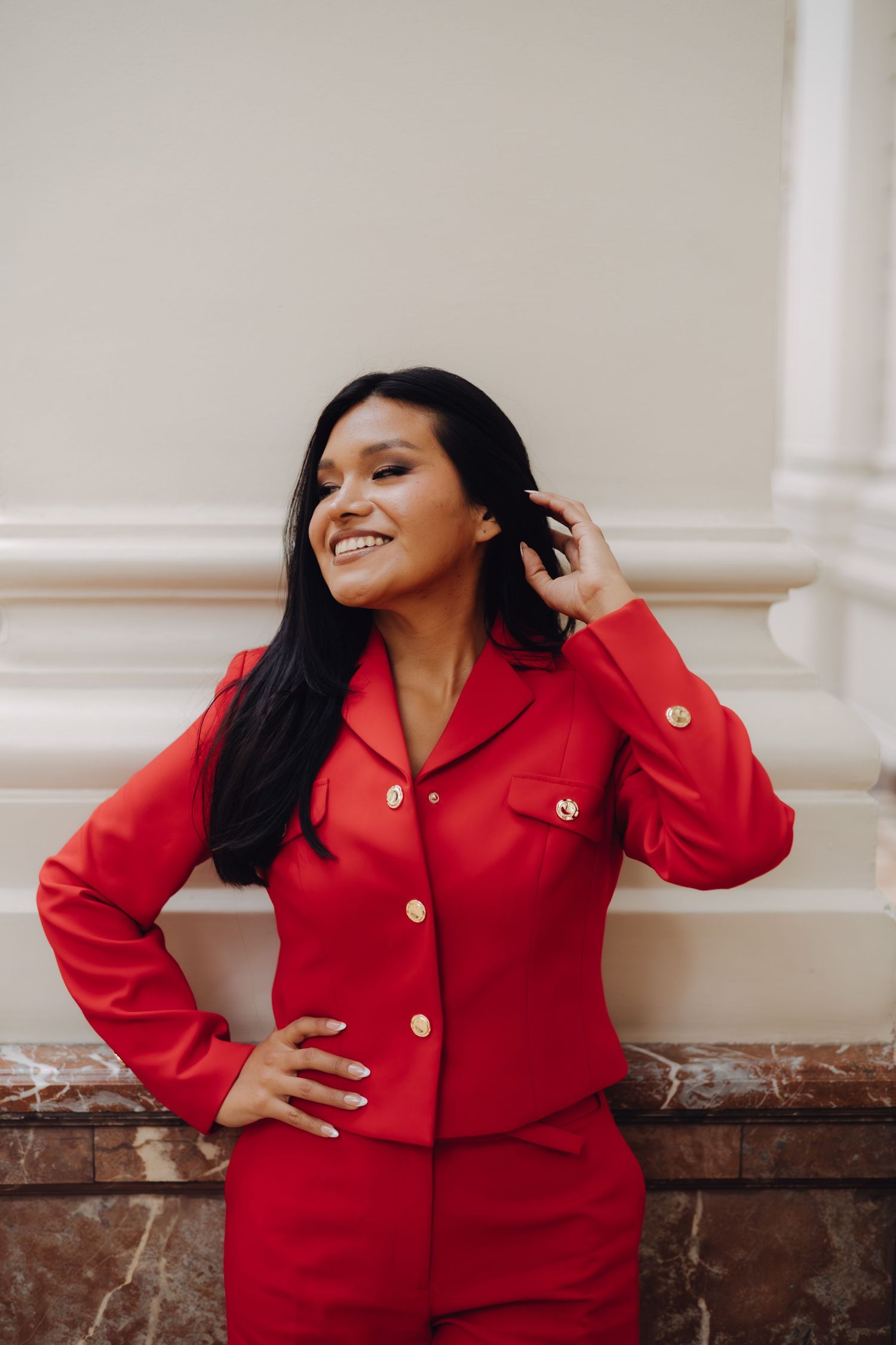Happy woman wearing a bold red suit with gold buttons, leaning against a beige wall outdoors.