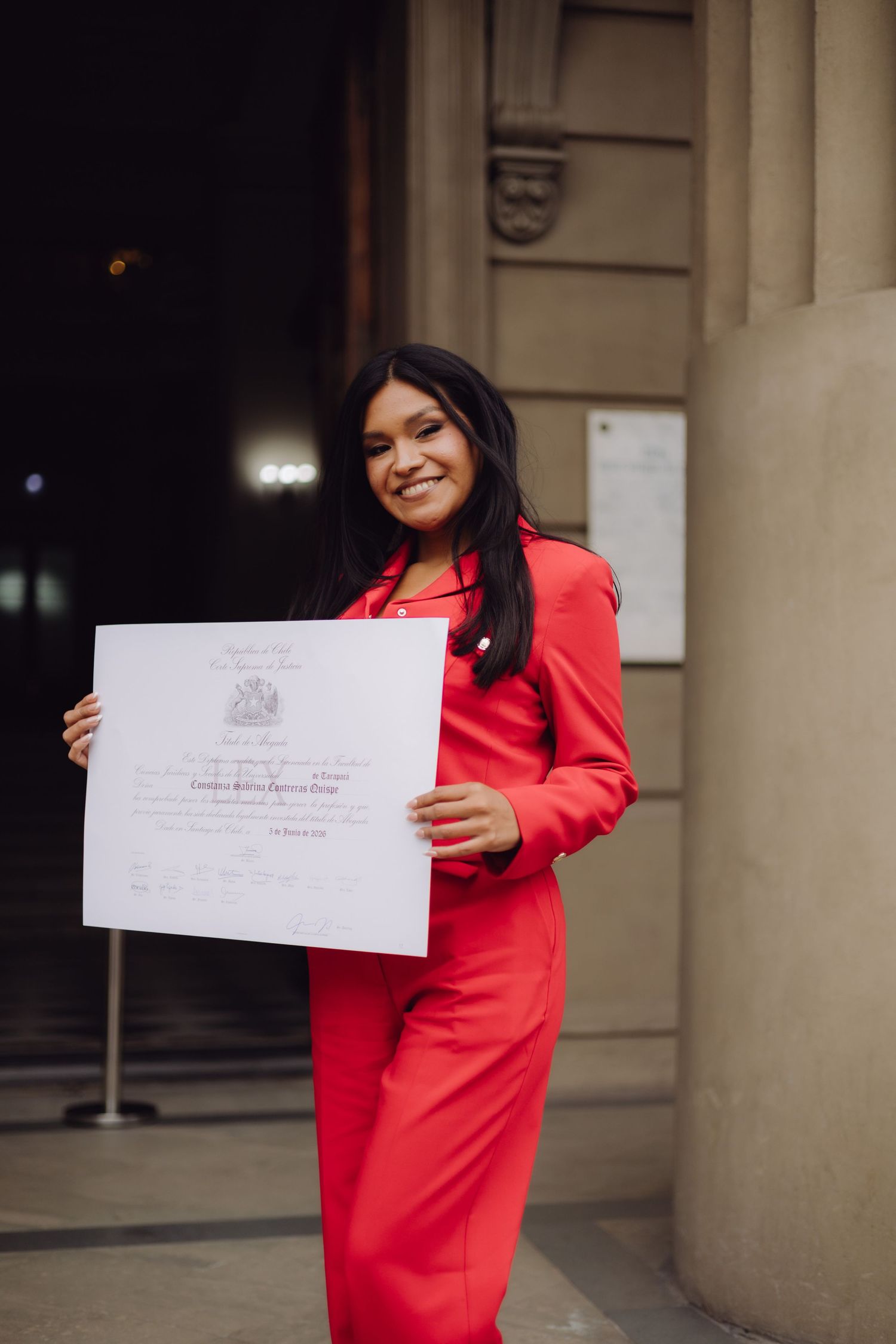 Happy woman in red suit proudly displaying an official certificate outside a historic building entrance.
