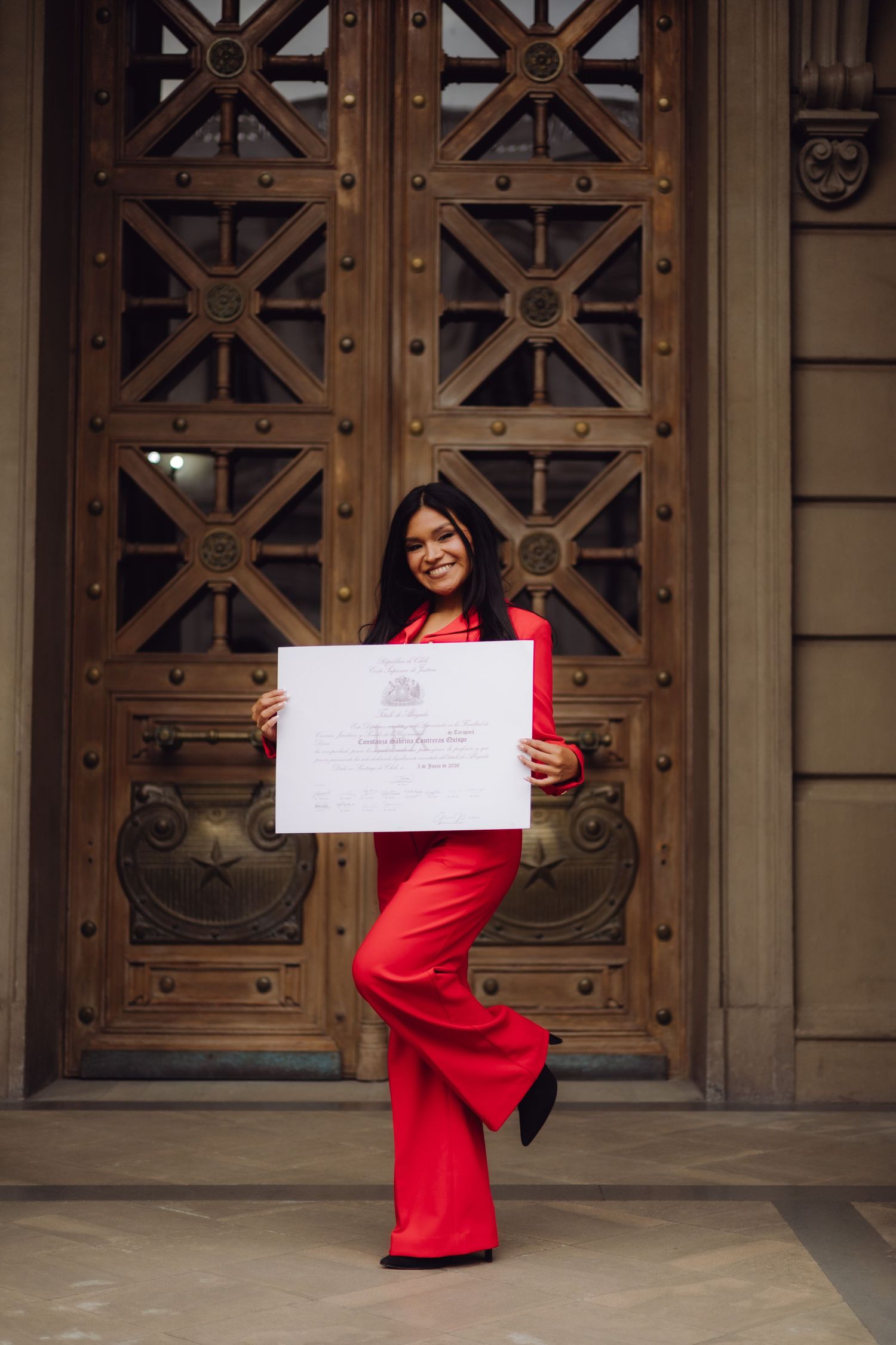 Joyful woman in red pantsuit holding an official certificate in front of ornate wooden doors.
