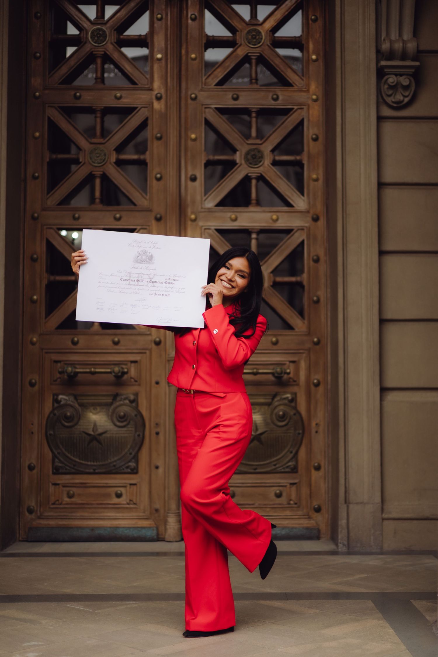 Excited woman in red suit posing playfully with an official certificate in front of ornate doors.
