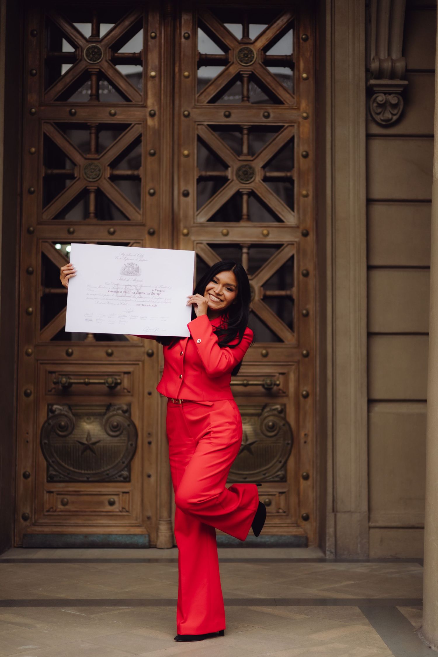 Cheerful woman in red outfit celebrating with an official certificate outside ornate wooden doors.