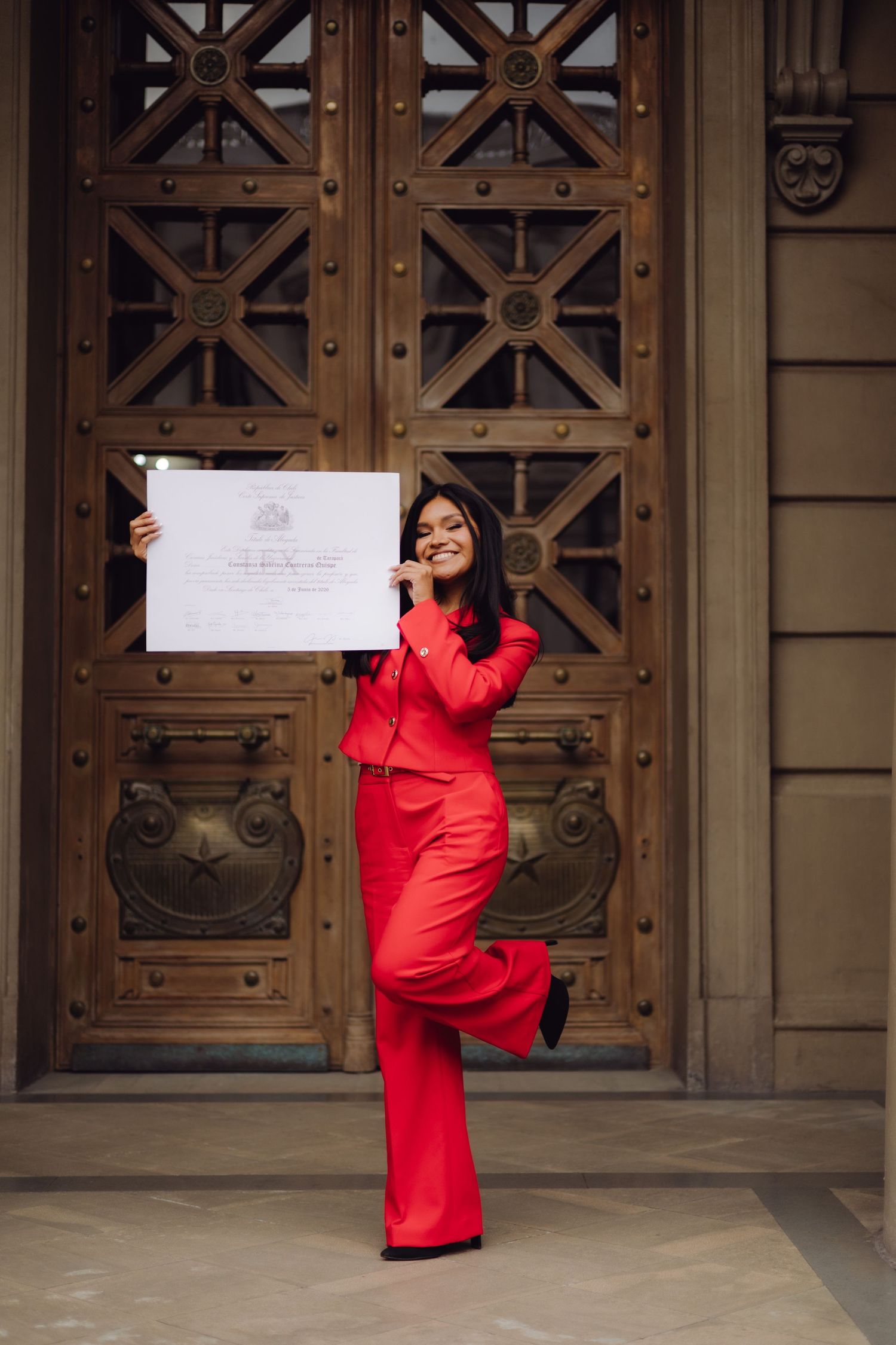 Smiling woman in red suit posing with an official certificate in front of decorative wooden doors.