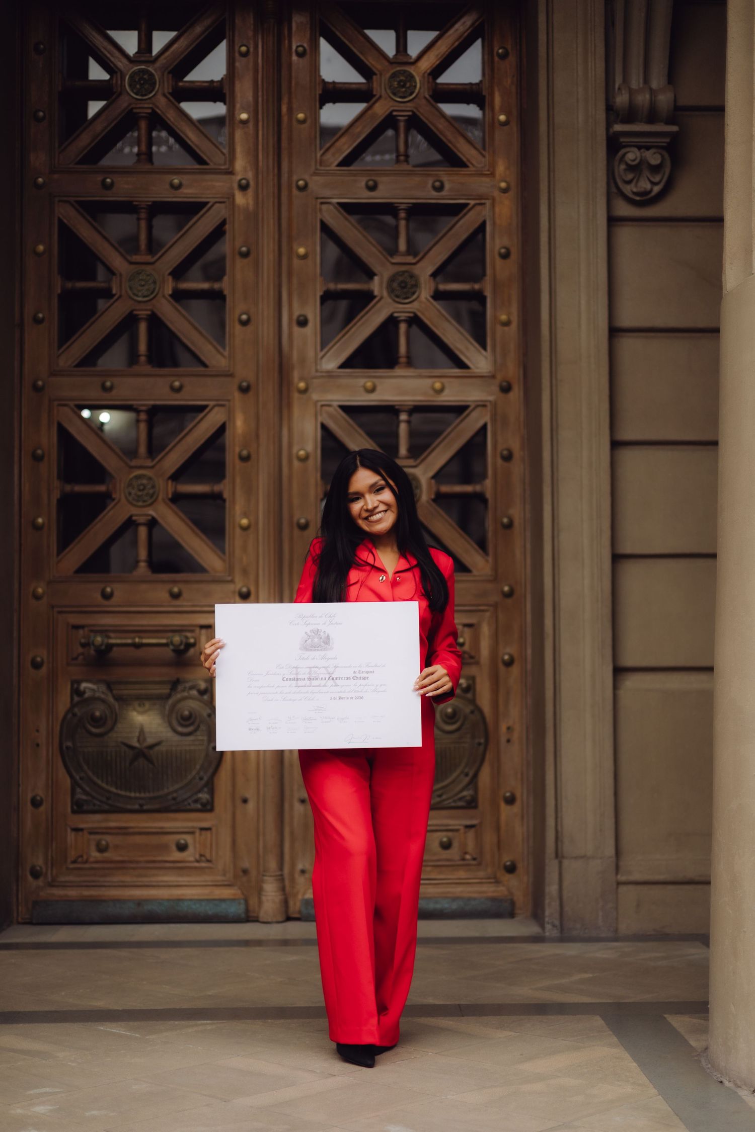Woman in red pantsuit proudly holding an official certificate while standing before large wooden doors.
